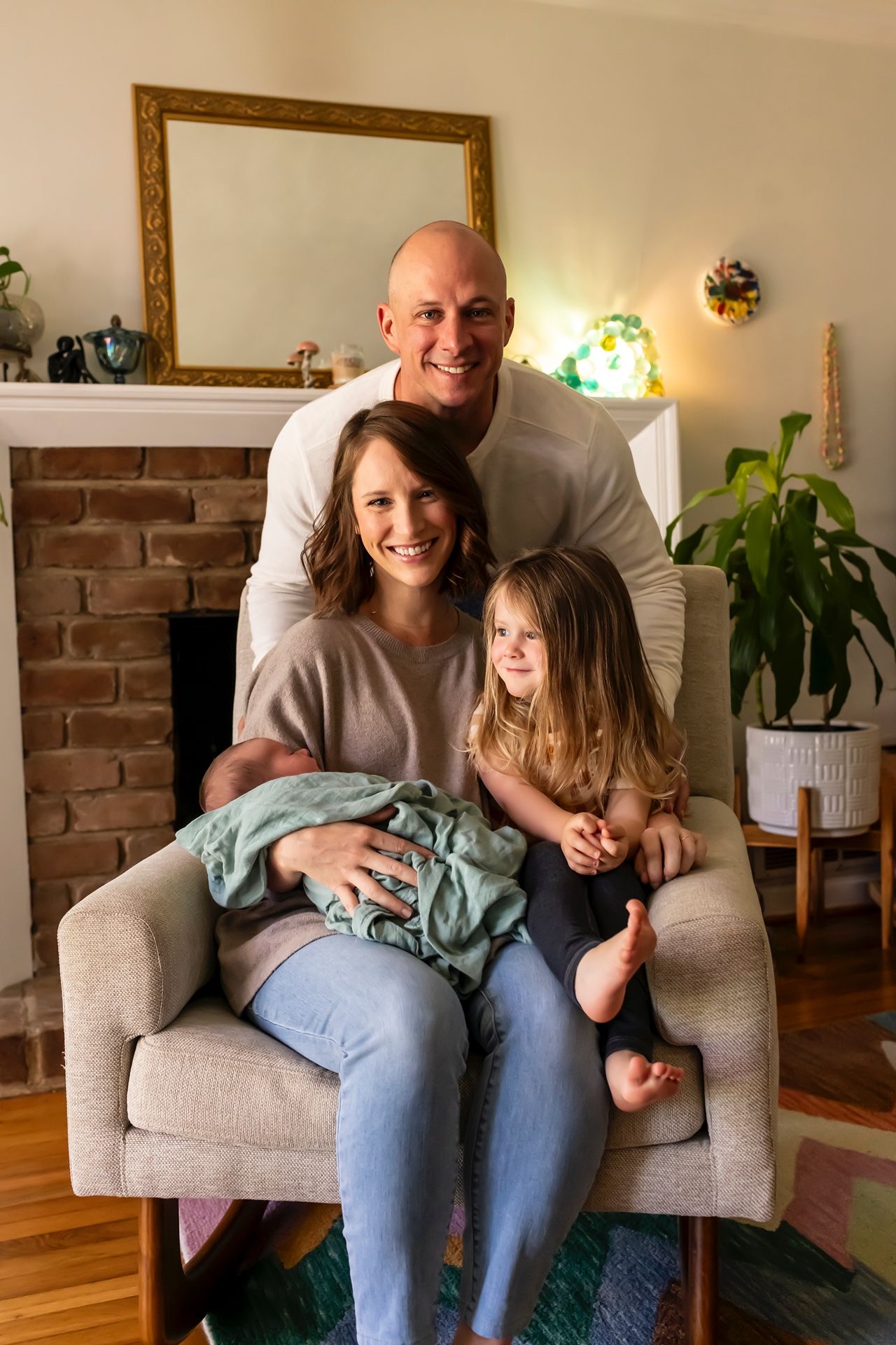 Family portrait in living room with brick fireplace, gathered in grey armchair.