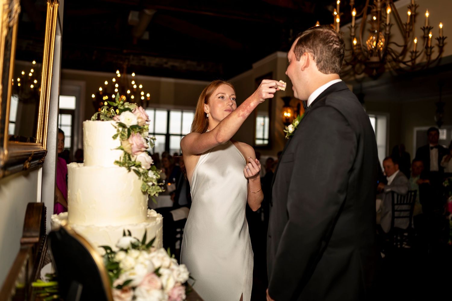 A couple participates in the traditional wedding cake feeding ritual during their reception under elegant chandeliers.