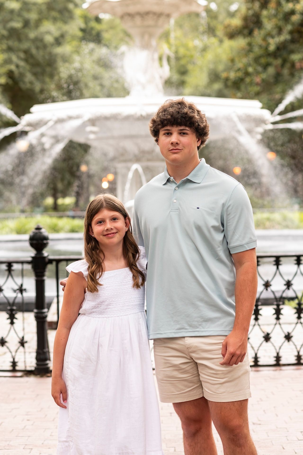 Siblings pose in front of a fountain wearing light colored clothing on a summer day.