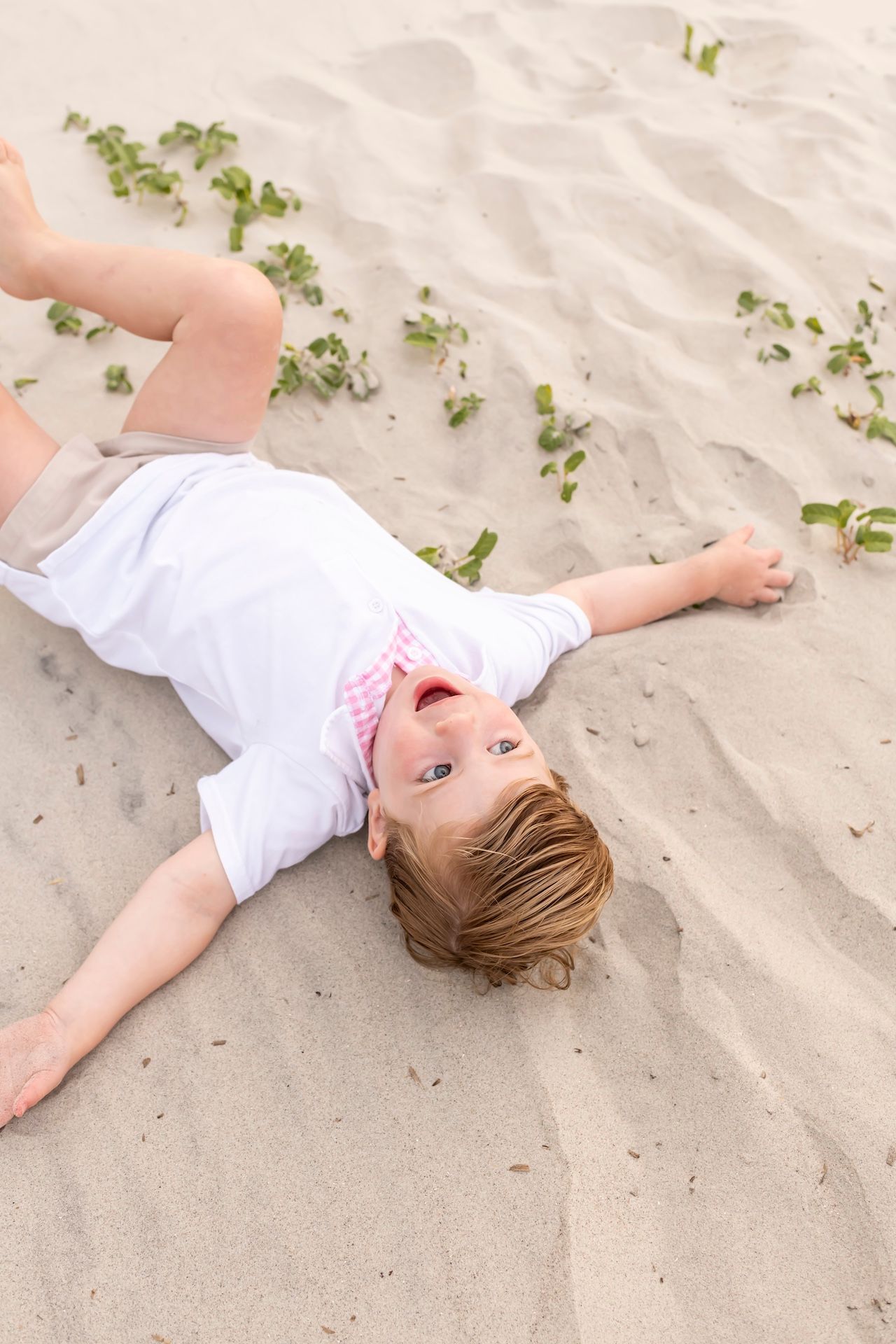 A young child lies on a sandy beach creating sand angels, wearing white clothing.