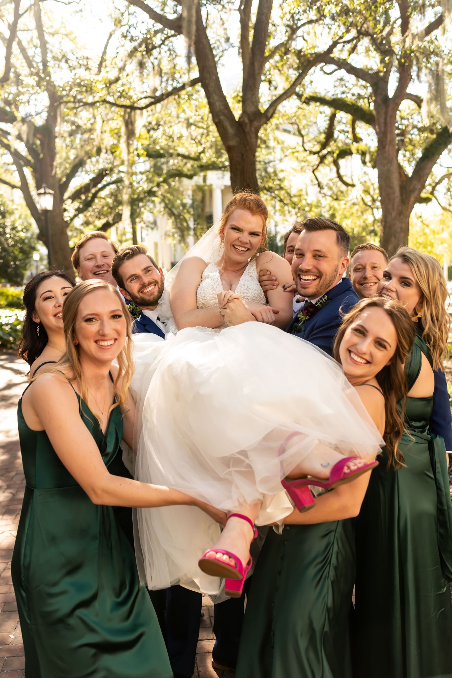 Wedding party in green bridesmaid dresses playfully lifting and holding up a bride in a white gown outdoors under oak trees.