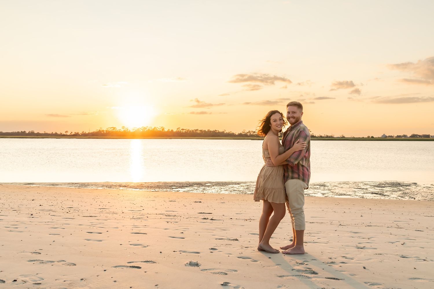 A couple embraces on a sandy beach at sunset with golden light reflecting off calm waters.