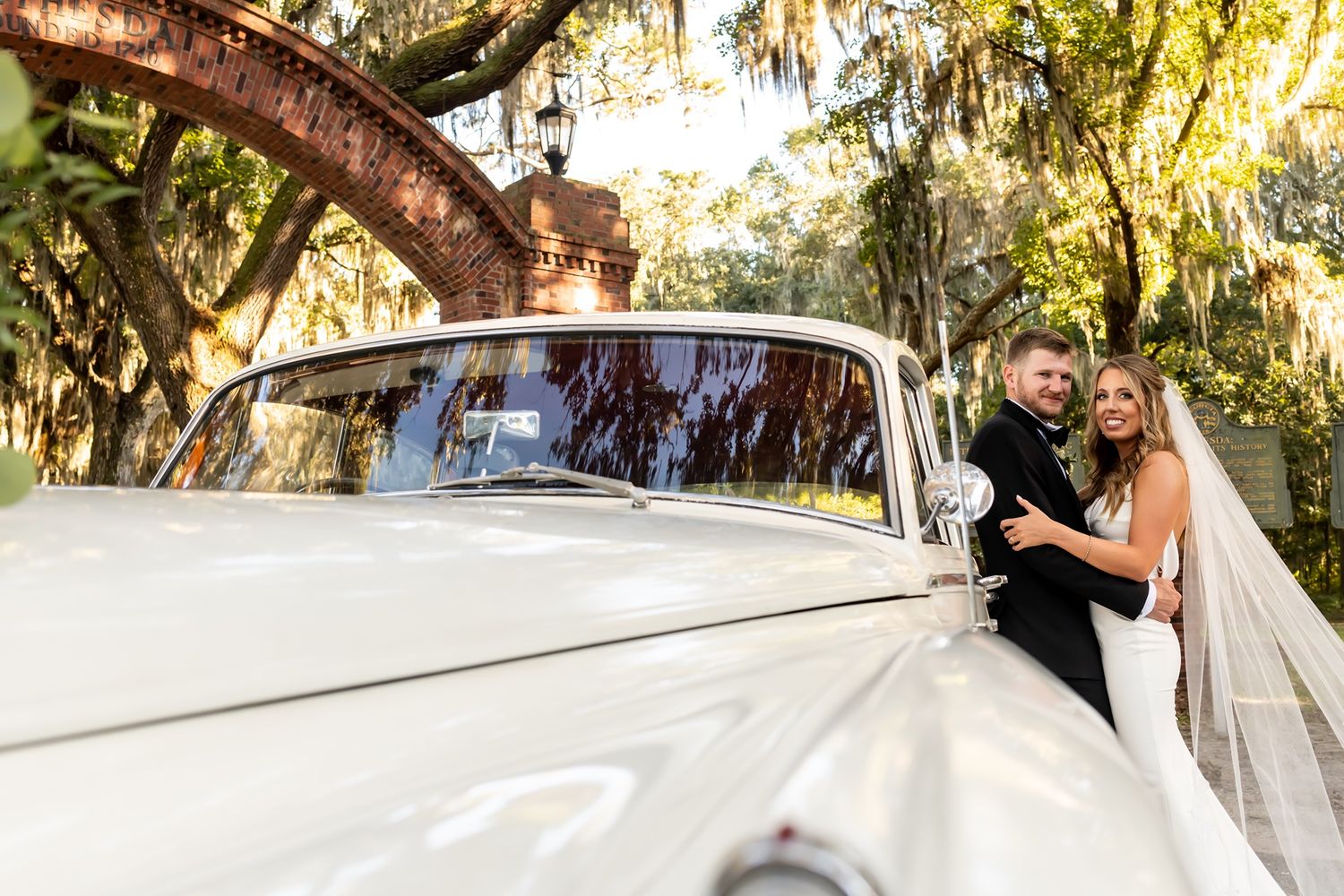 A classic white vintage car parked under moss-draped oak trees at a sunny outdoor wedding celebration.