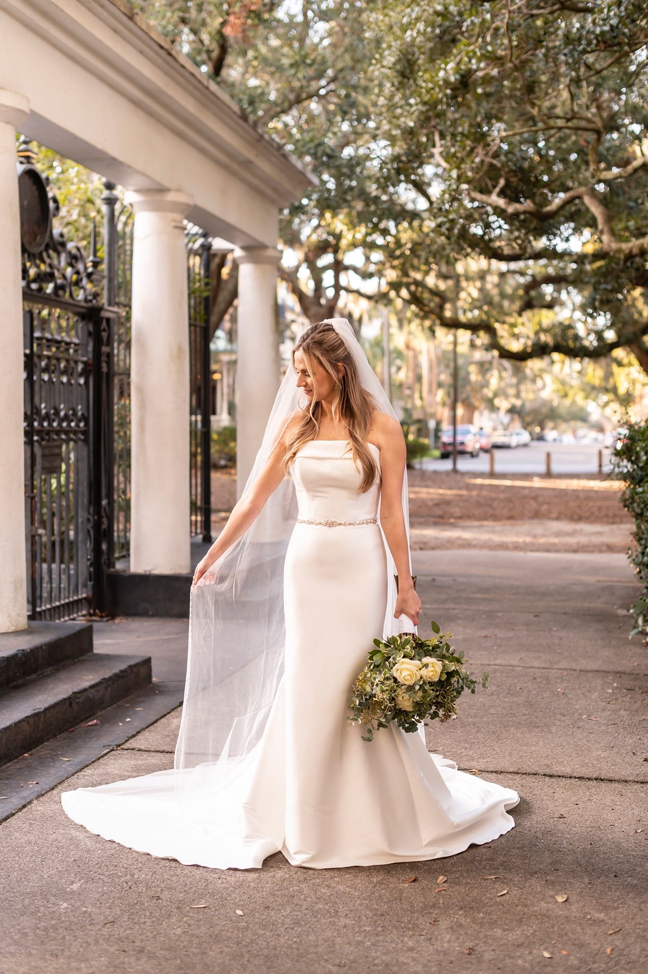 A bride in a flowing white gown poses elegantly among white columns and iron gates in a garden setting.