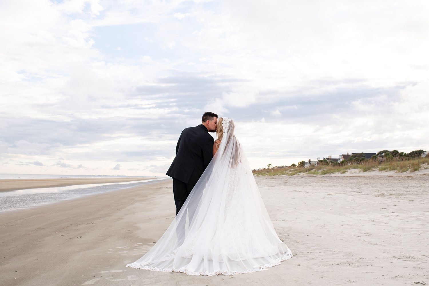 A flowing white wedding dress billows in the ocean breeze as figures walk along a sandy beach at sunset.