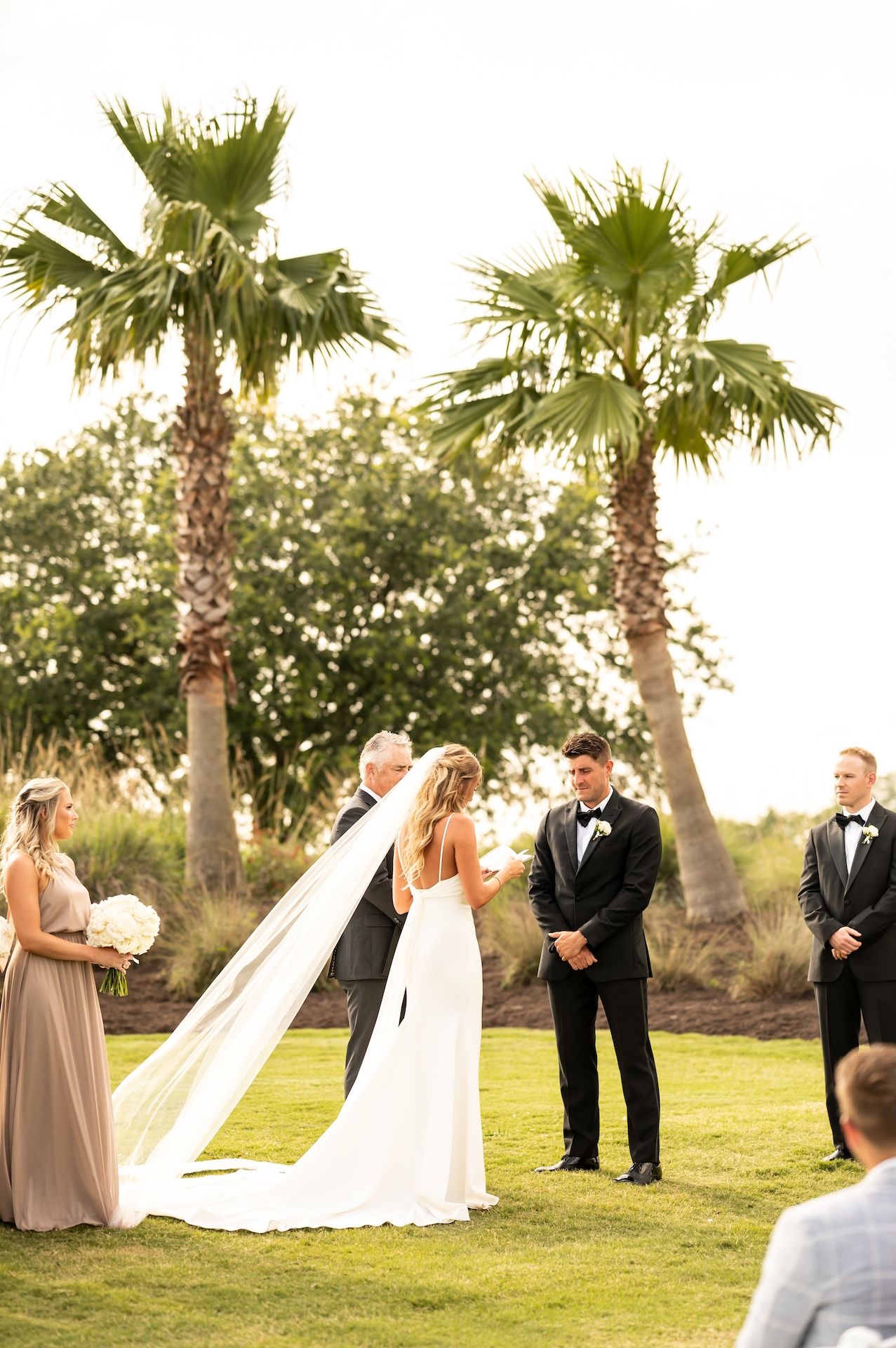 Outdoor wedding ceremony on green lawn with palm trees, bride in white gown with veil, groom in black tuxedo.