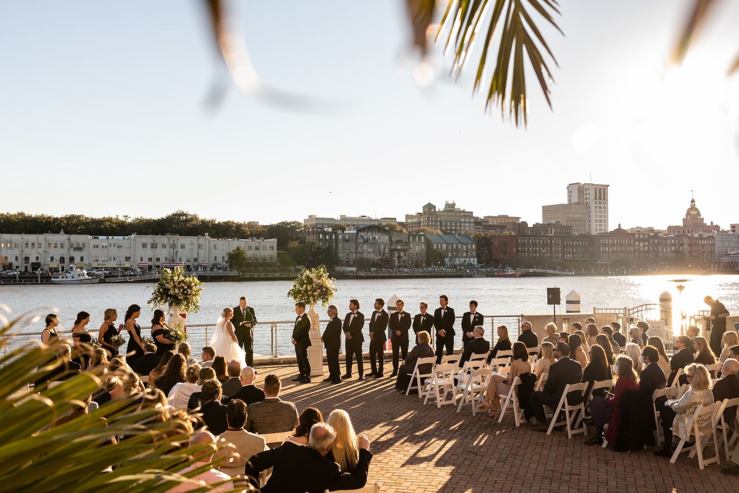 Outdoor waterfront wedding ceremony at sunset with guests seated in white chairs and a city skyline backdrop.