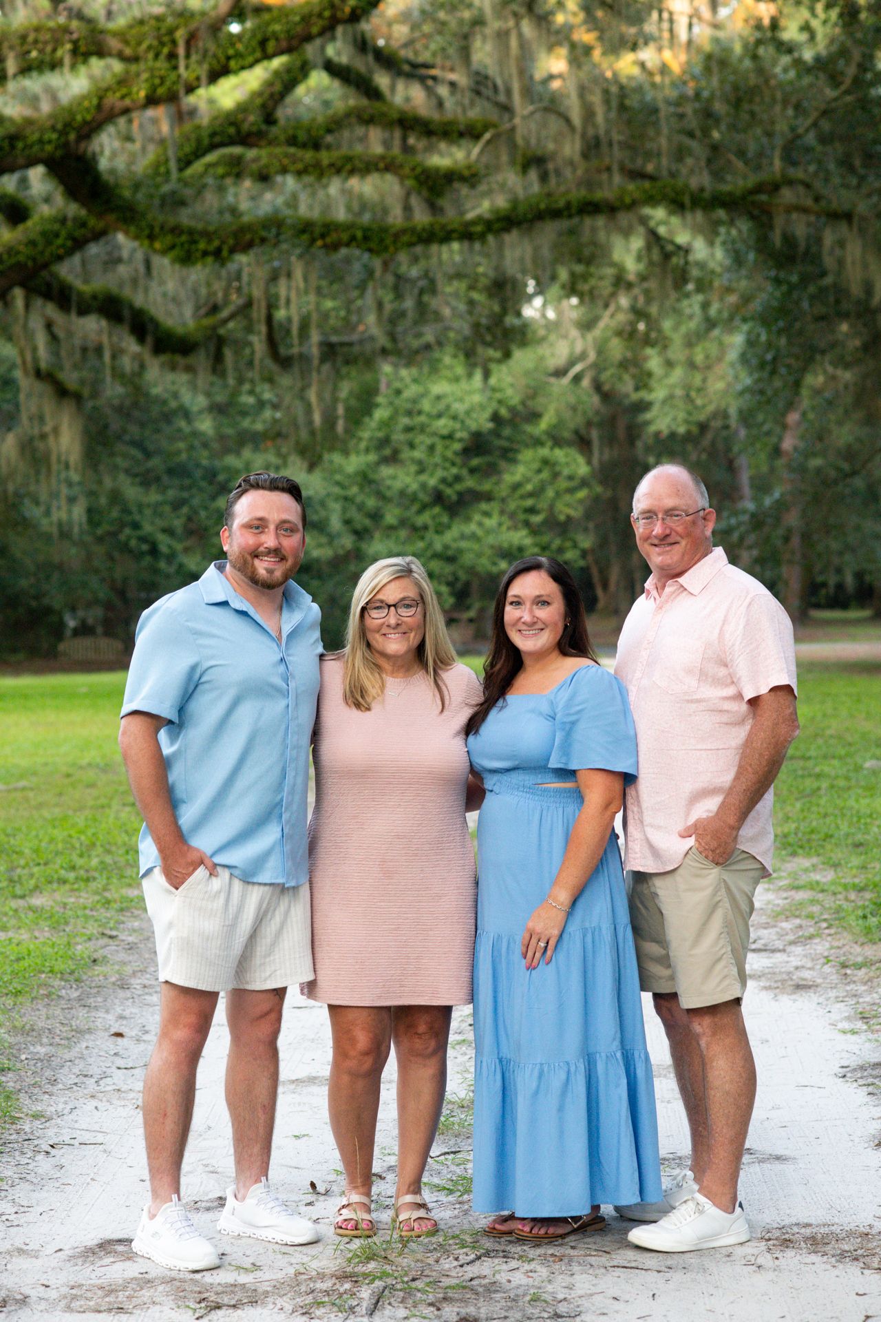 A family group wearing coordinated blue, pink and white outfits poses together outdoors under moss-draped trees.