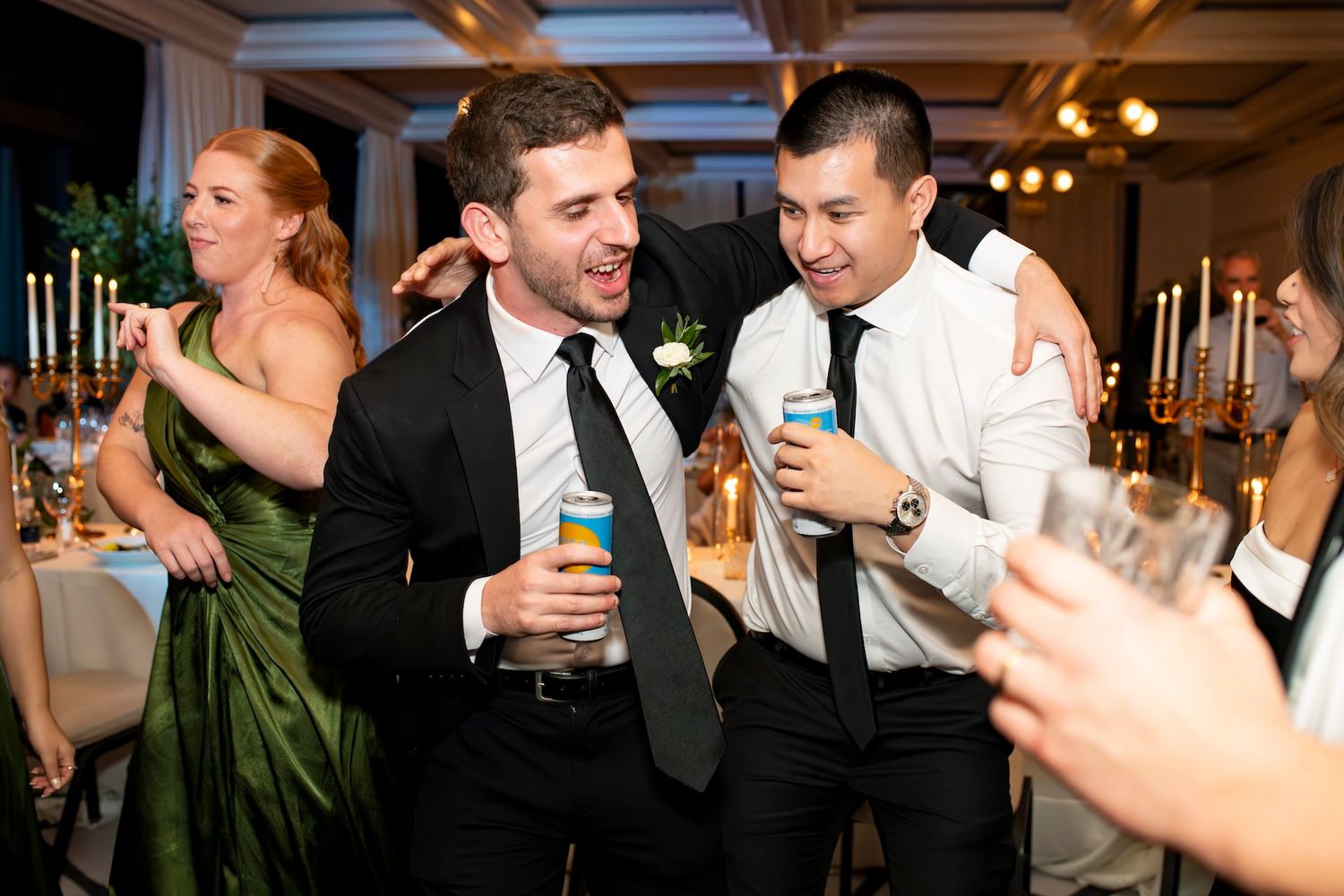 Wedding guests laugh and dance together while holding drinks at an energetic reception party.