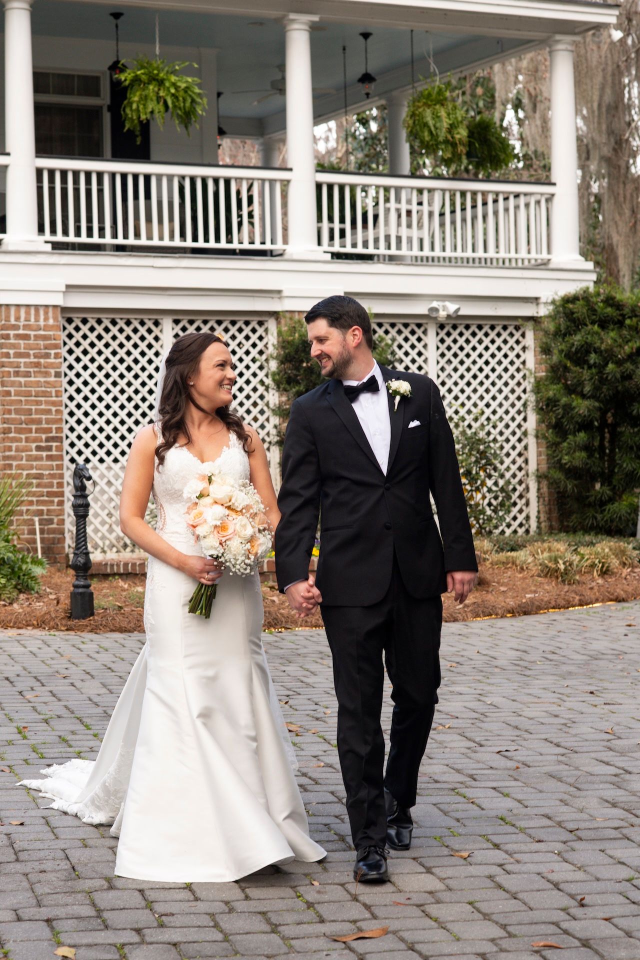 Bride in white gown and groom in black tuxedo walk hand in hand on cobblestone path before elegant white plantation home.