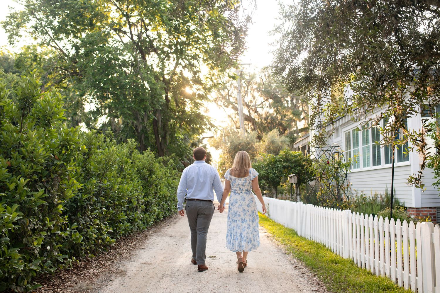 People walk along a sunlit garden path bordered by lush greenery and a white picket fence.