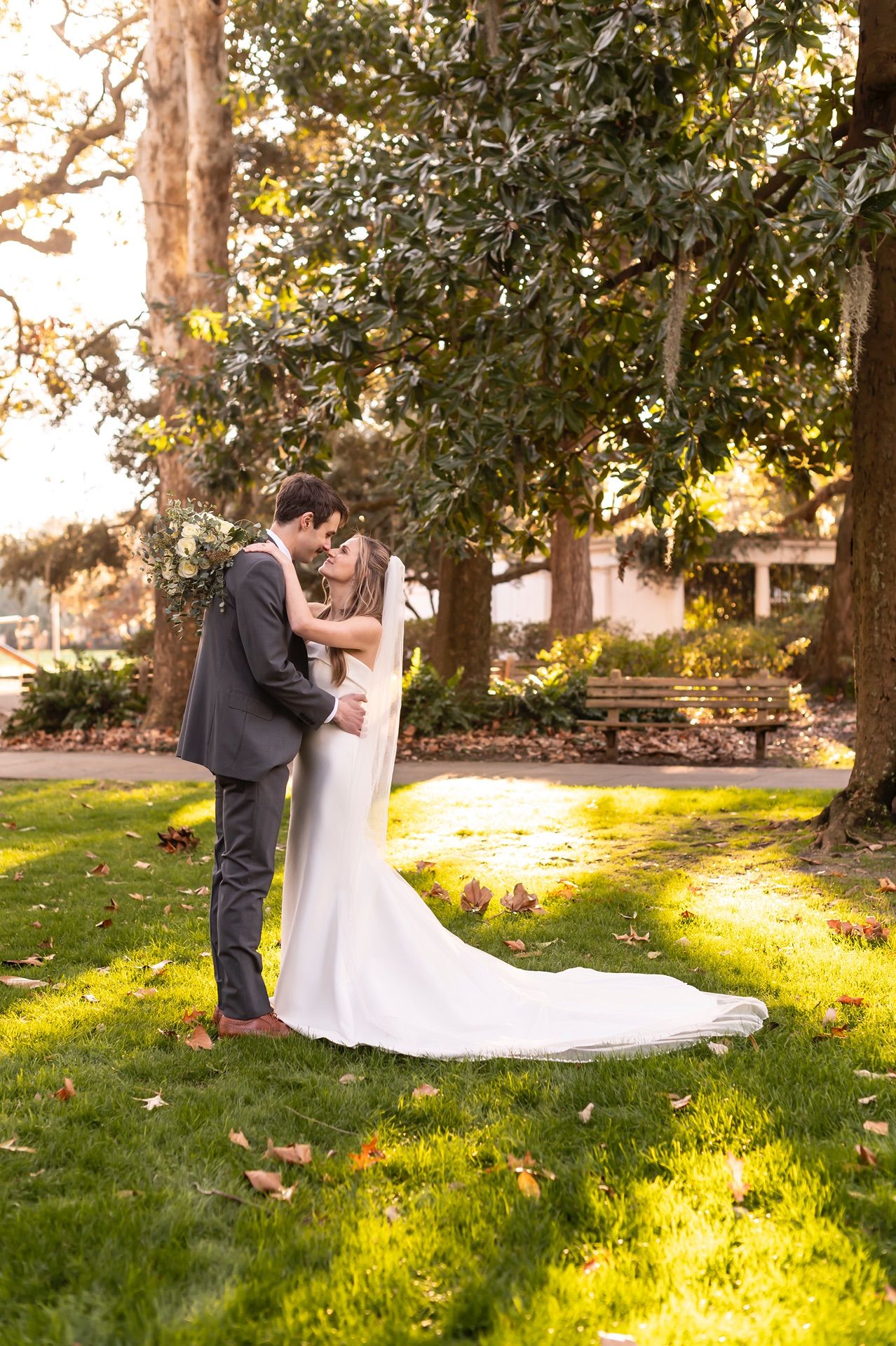 A couple shares a romantic sunset moment on a grass lawn beneath mature trees with golden sunlight filtering through.