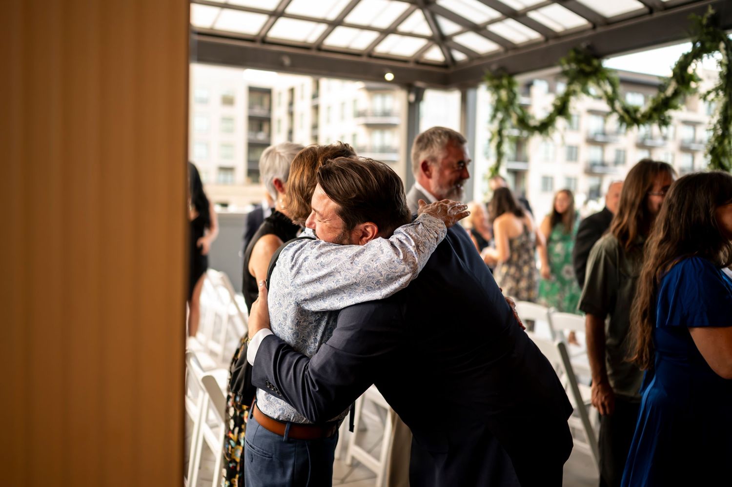 Wedding reception venue with guests hugging and mingling under skylight and indoor greenery.