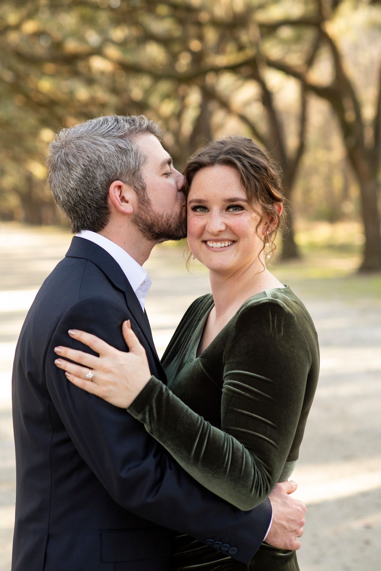 A couple in formal attire embrace outdoors, with the woman wearing a green velvet top and the man in a dark suit.
