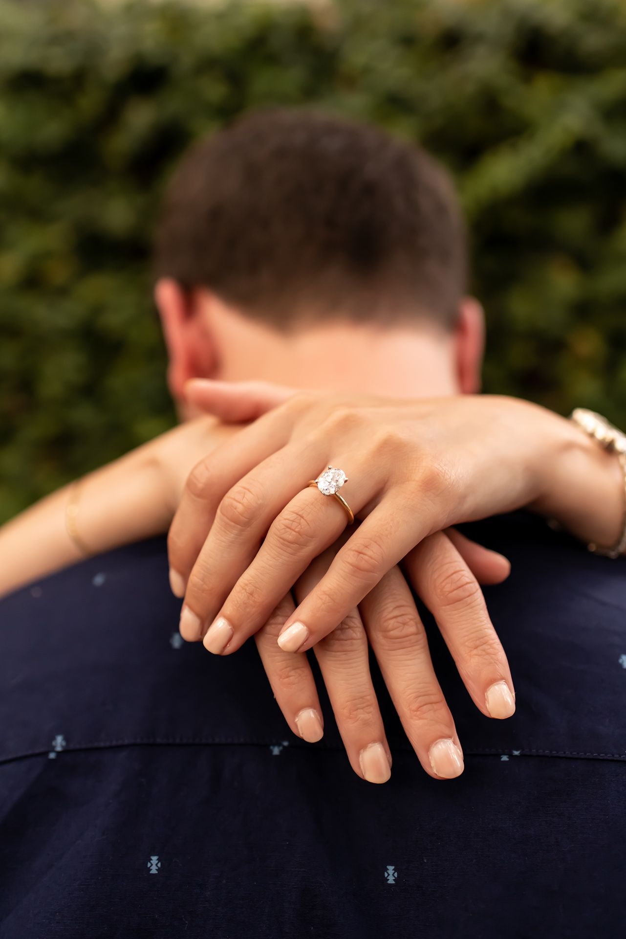 Close-up of hands displaying an engagement ring with a sparkling diamond solitaire on a navy blue background.