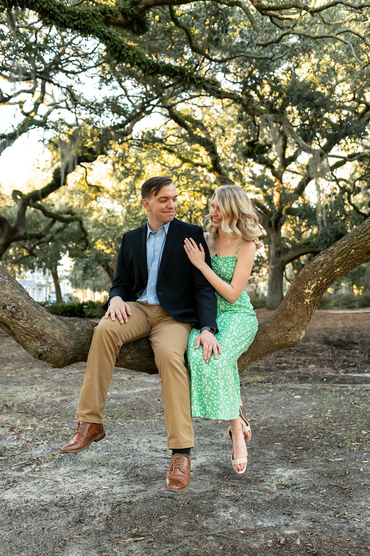 A couple shares intimate moments while sitting on a large tree branch in a natural outdoor setting during sunset.