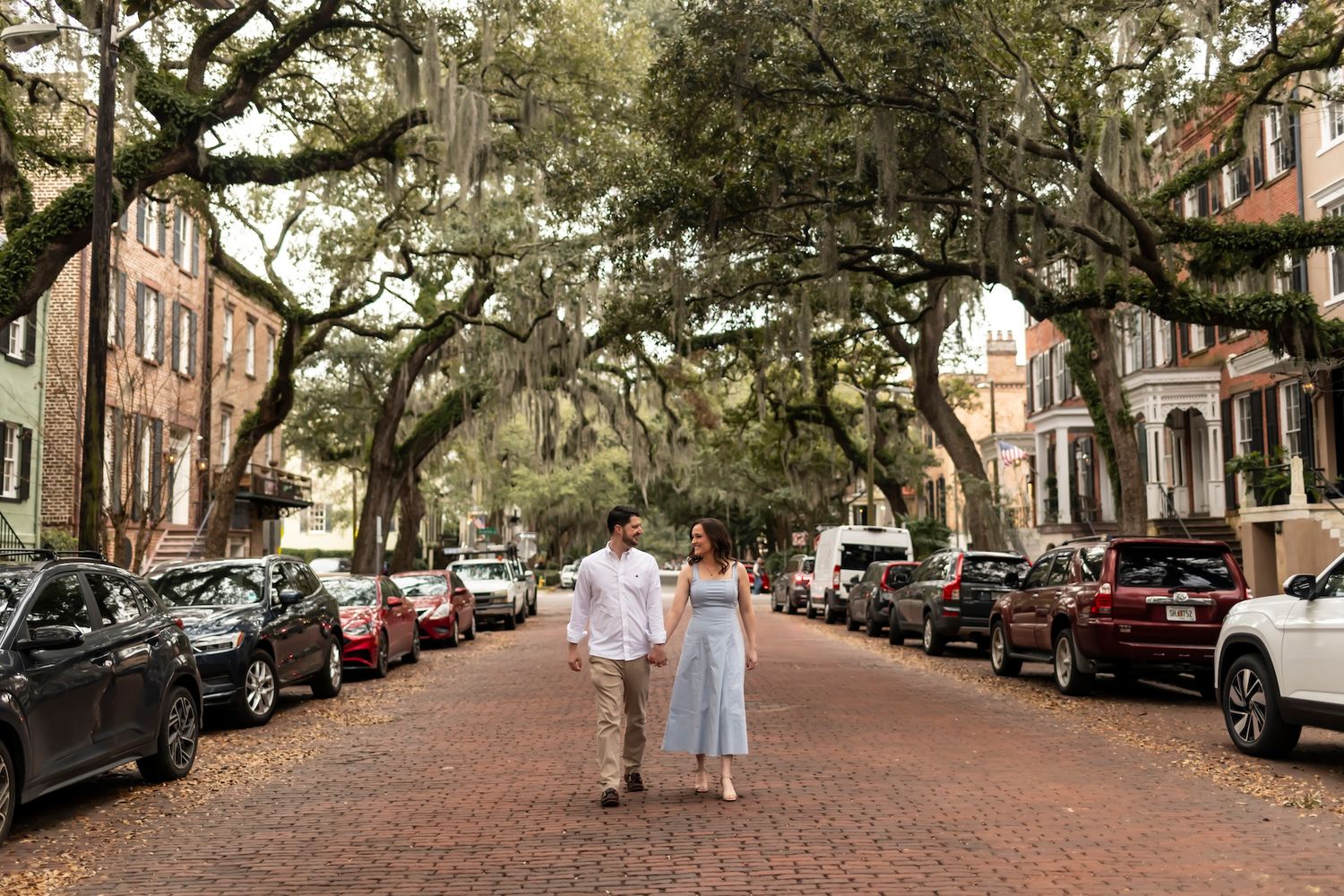 A couple strolls down a romantic brick-paved street lined with Spanish moss-draped oak trees in historic Savannah, Georgia.