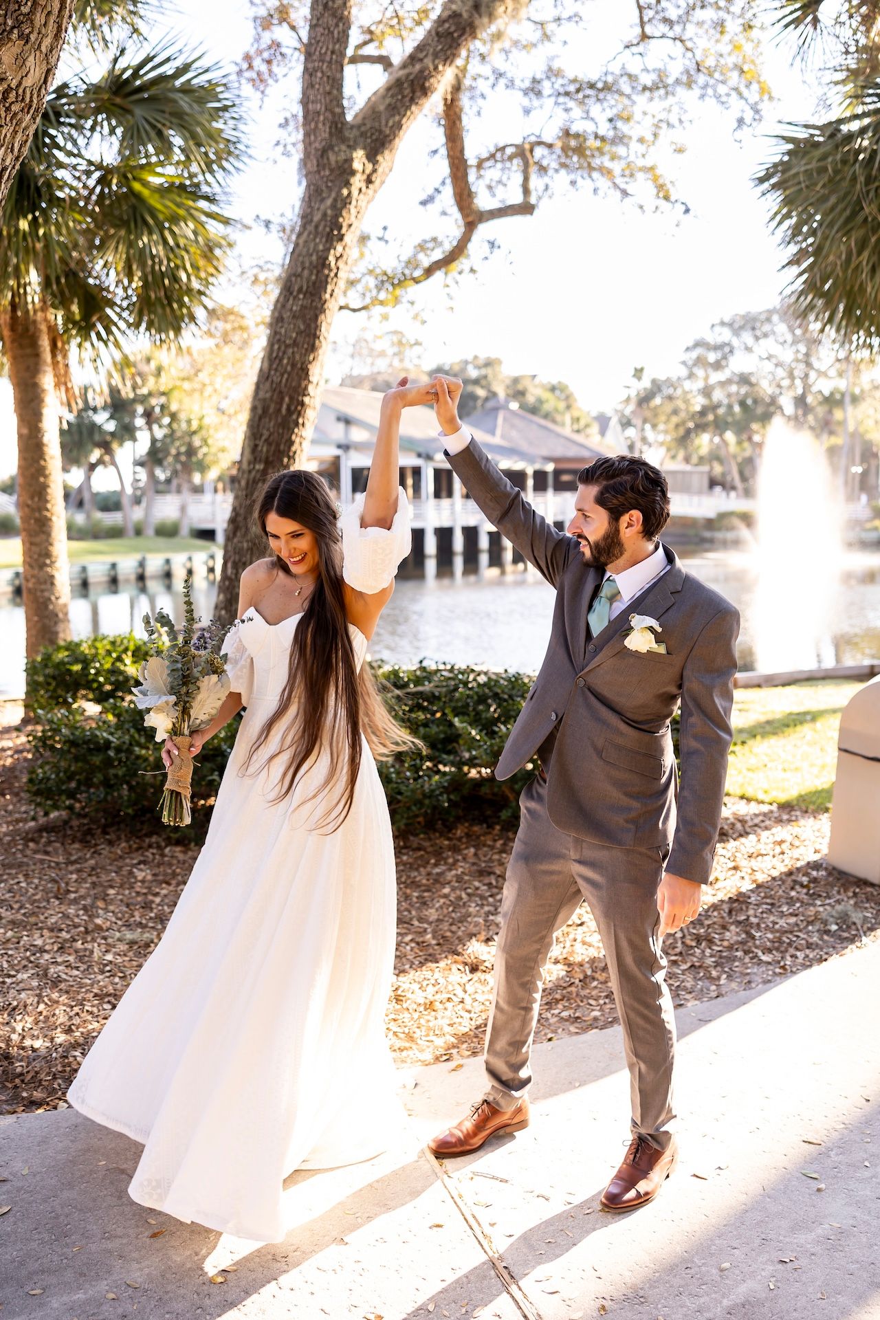 A joyful couple dances together outdoors near a lake at sunset, with palm trees and natural light surrounding them.