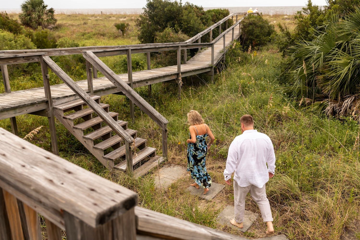 A wooden boardwalk with stairs winds through coastal dunes and vegetation as two people descend the steps.
