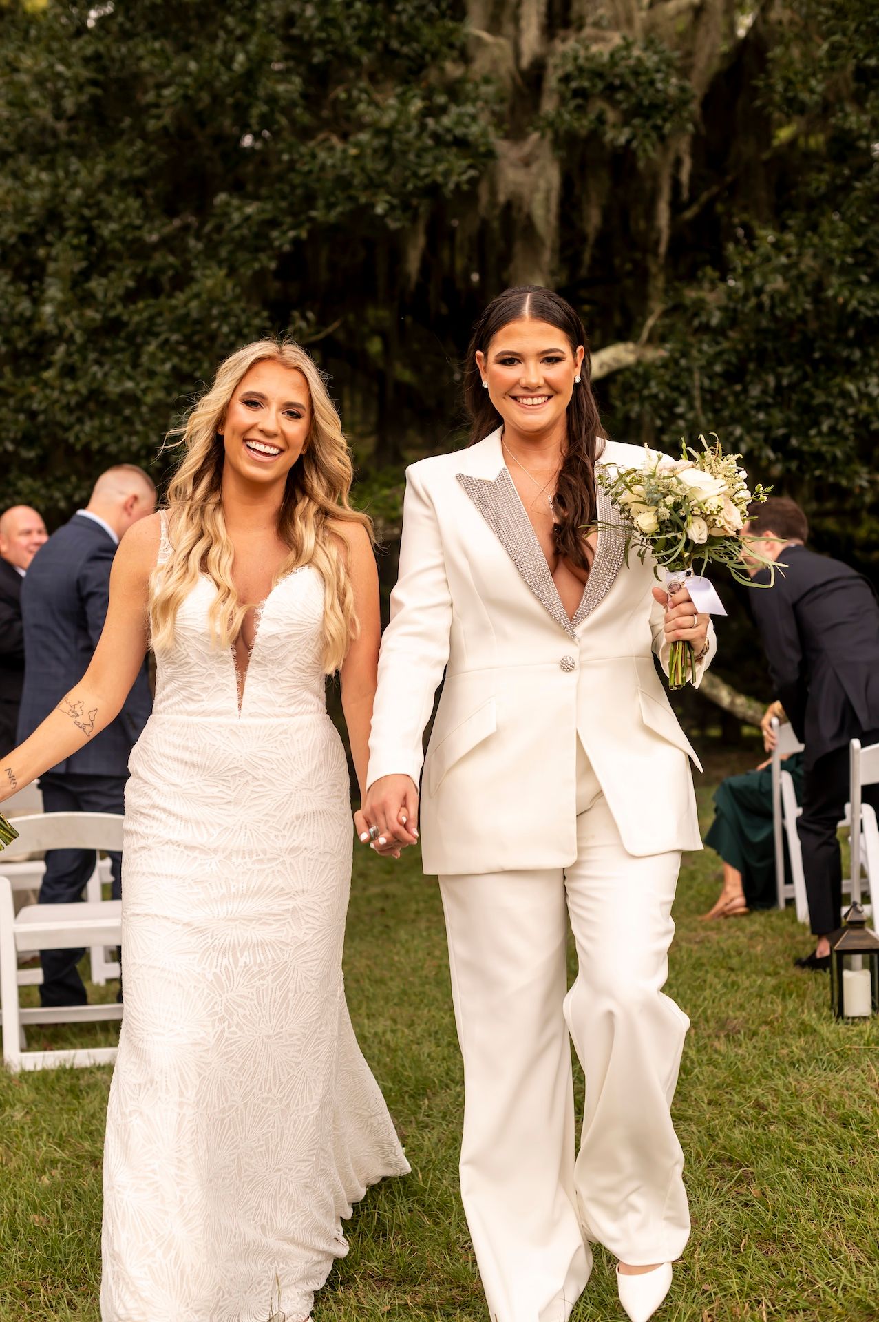 A couple in bridal attire hold hands on their wedding day, one in a white suit with bouquet and one in lace gown.