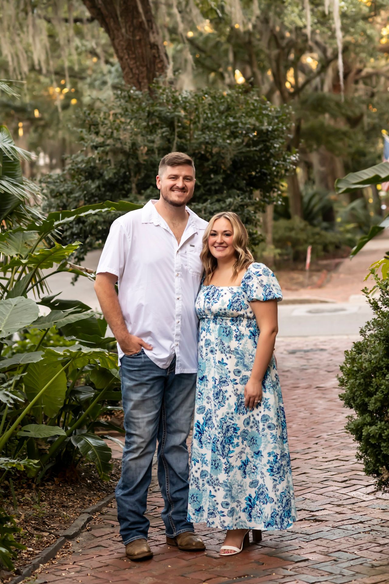 A couple stands on a brick pathway surrounded by lush tropical foliage and Spanish moss draped trees.