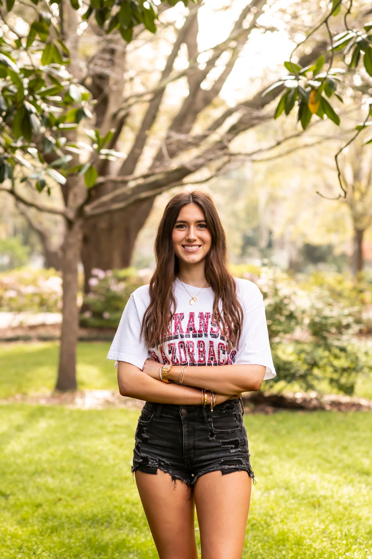 A person wearing a white t-shirt and denim shorts poses casually in front of trees on a sunny spring day in a park setting.