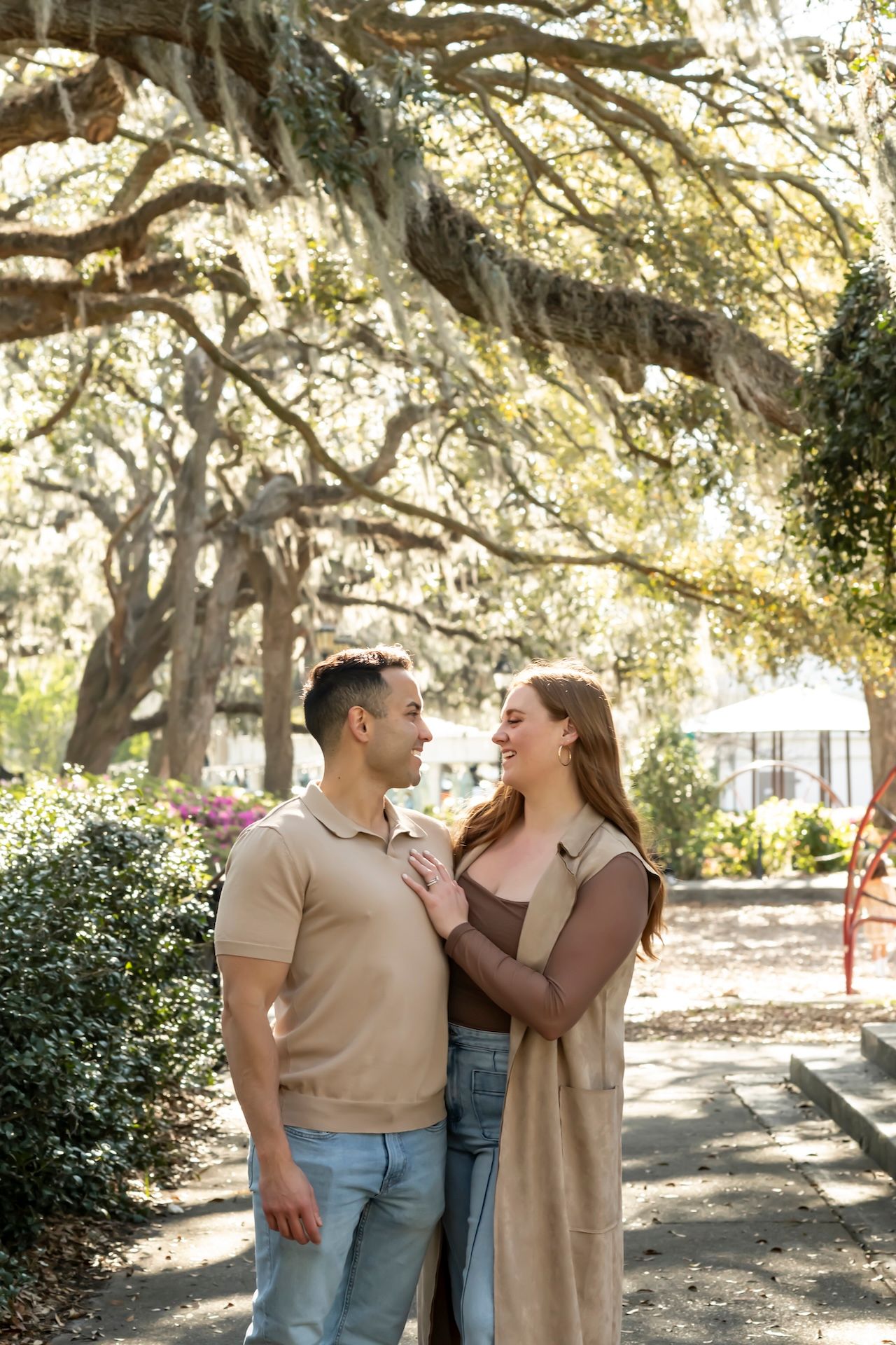 A couple shares a joyful moment together while standing on a path beneath Spanish moss-draped trees in a park setting.