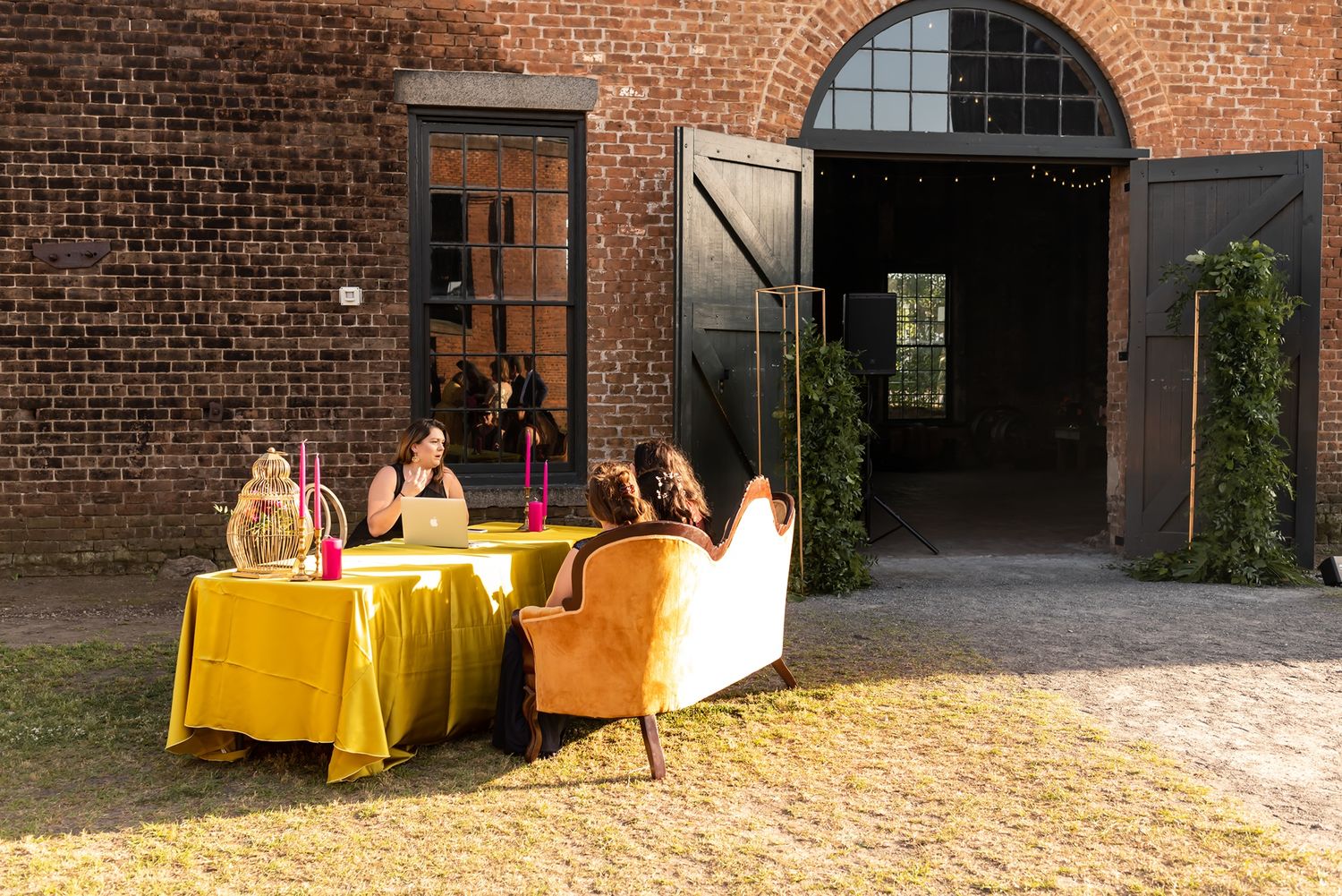 People gather at yellow-clothed tables outside a rustic brick building entrance on a sunny afternoon.
