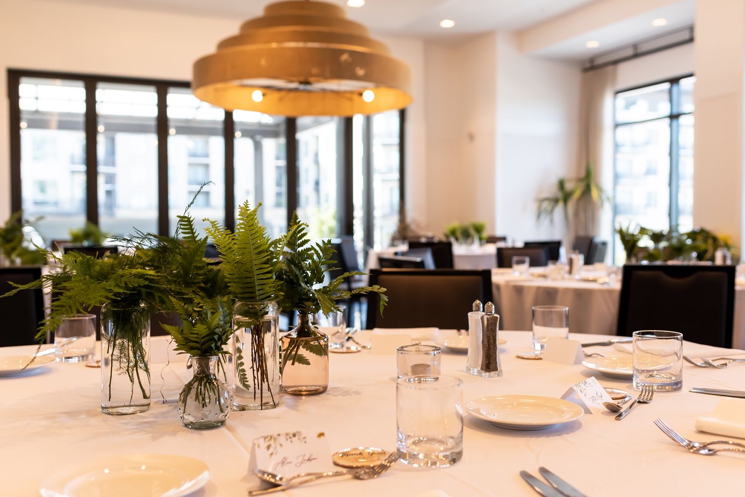 Modern restaurant interior with long white tables, elegant place settings and natural lighting illuminating the dining space.