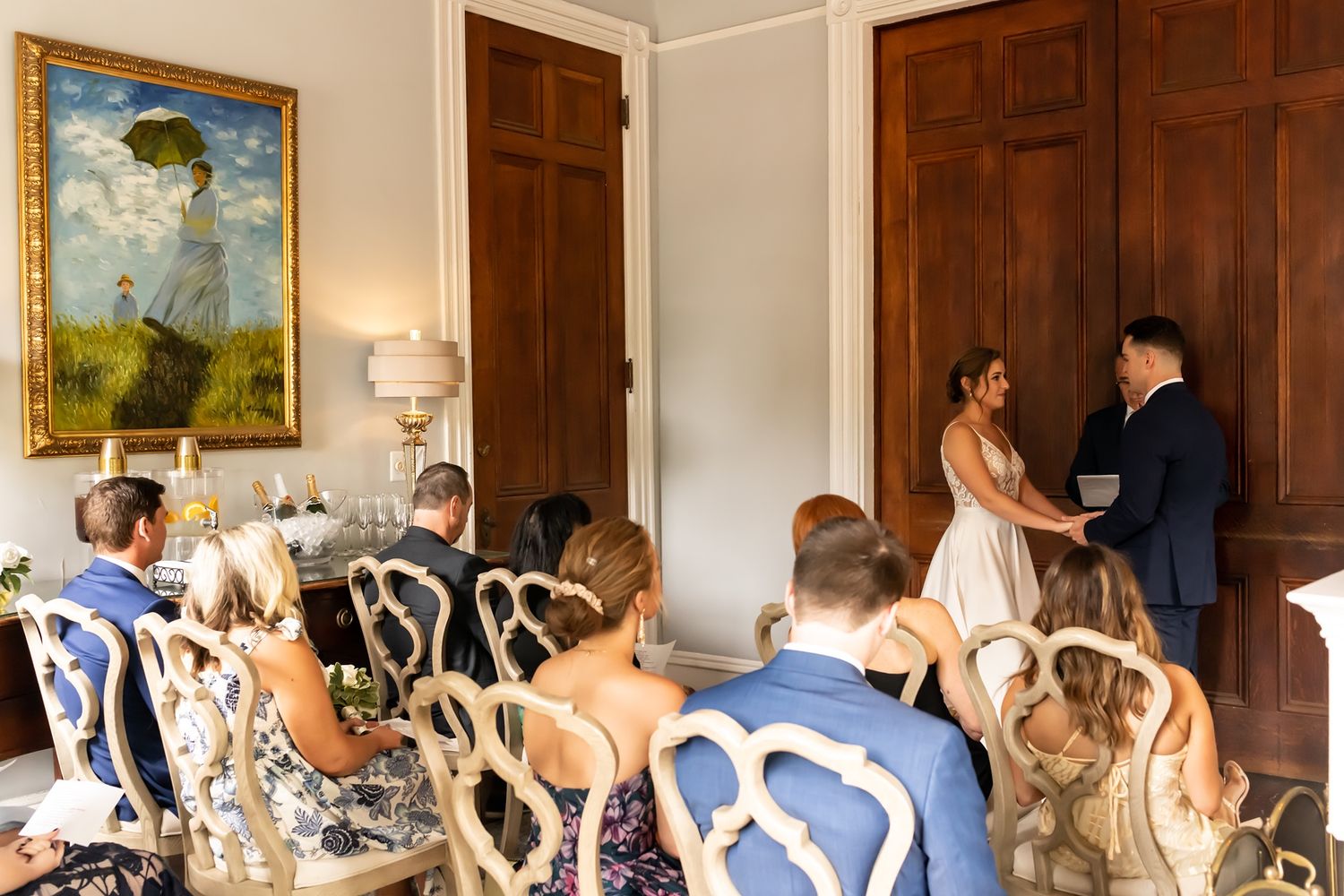Wedding guests seated on ornate chairs watching ceremony in front of vintage art and wood paneling.