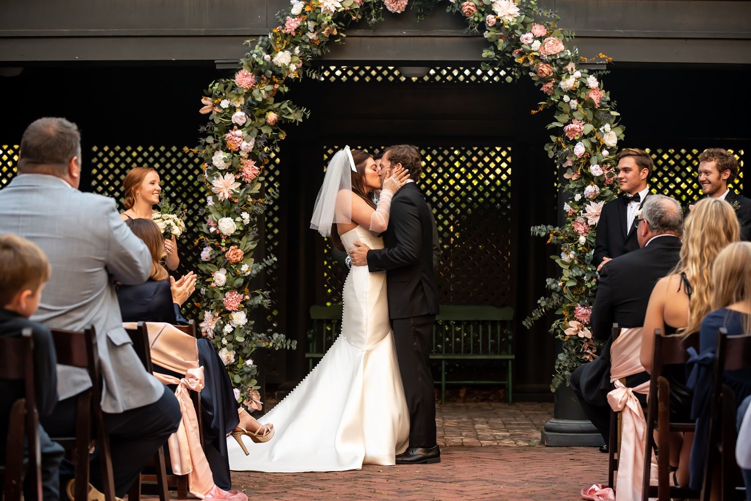 A couple shares a romantic kiss under a floral arch during an intimate garden wedding ceremony with guests looking on.