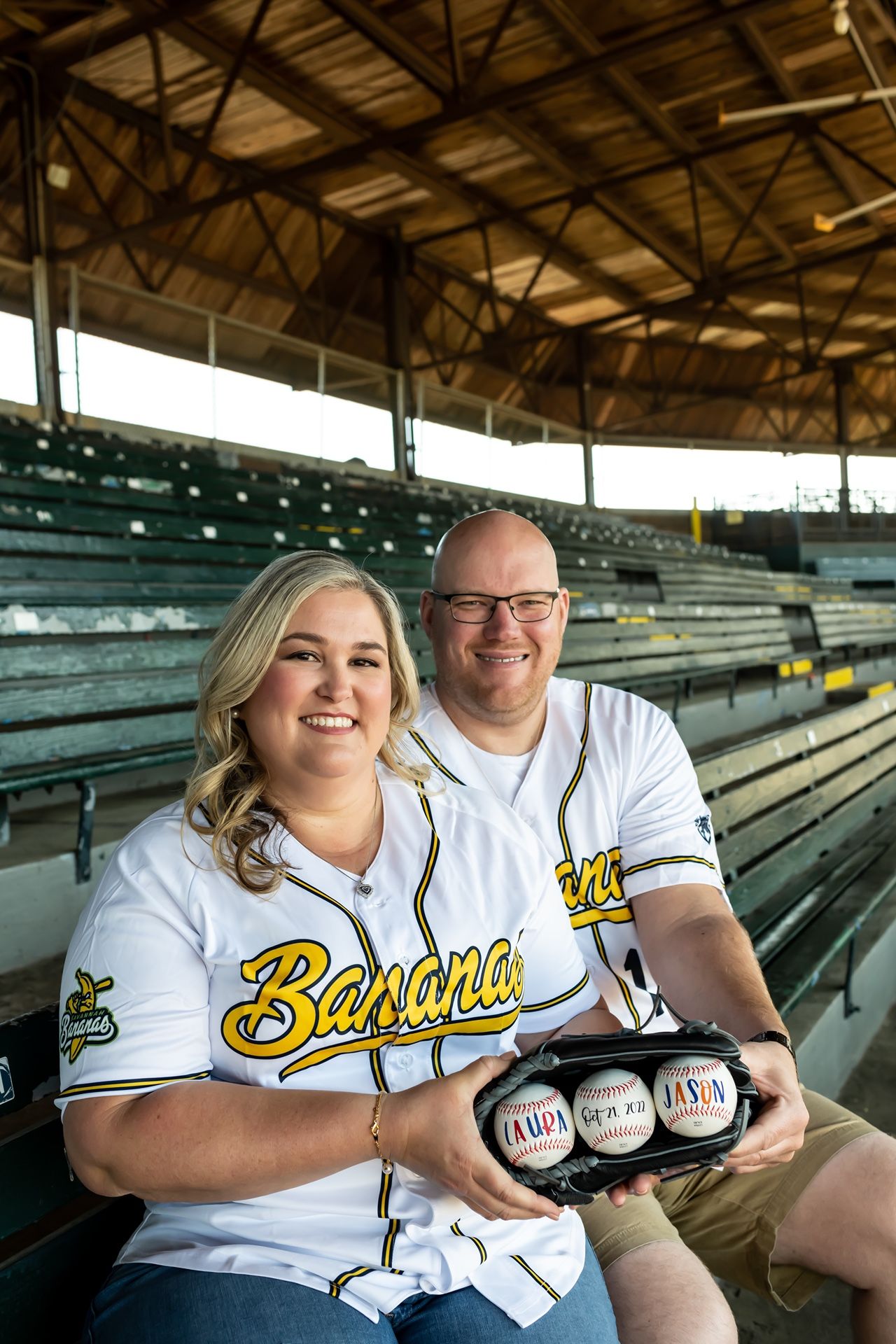 A couple in matching baseball jerseys holds beverages while sitting in empty stadium bleachers.