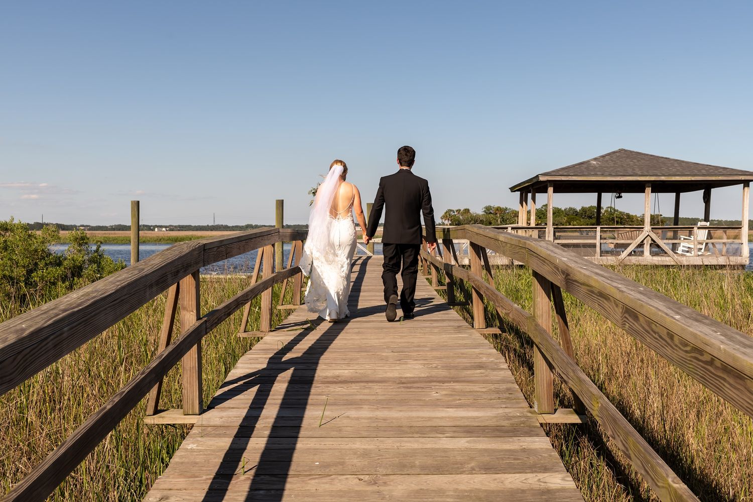 Wedding couple walks down a wooden boardwalk walkway along a beach with sunset shadows stretching across the path.