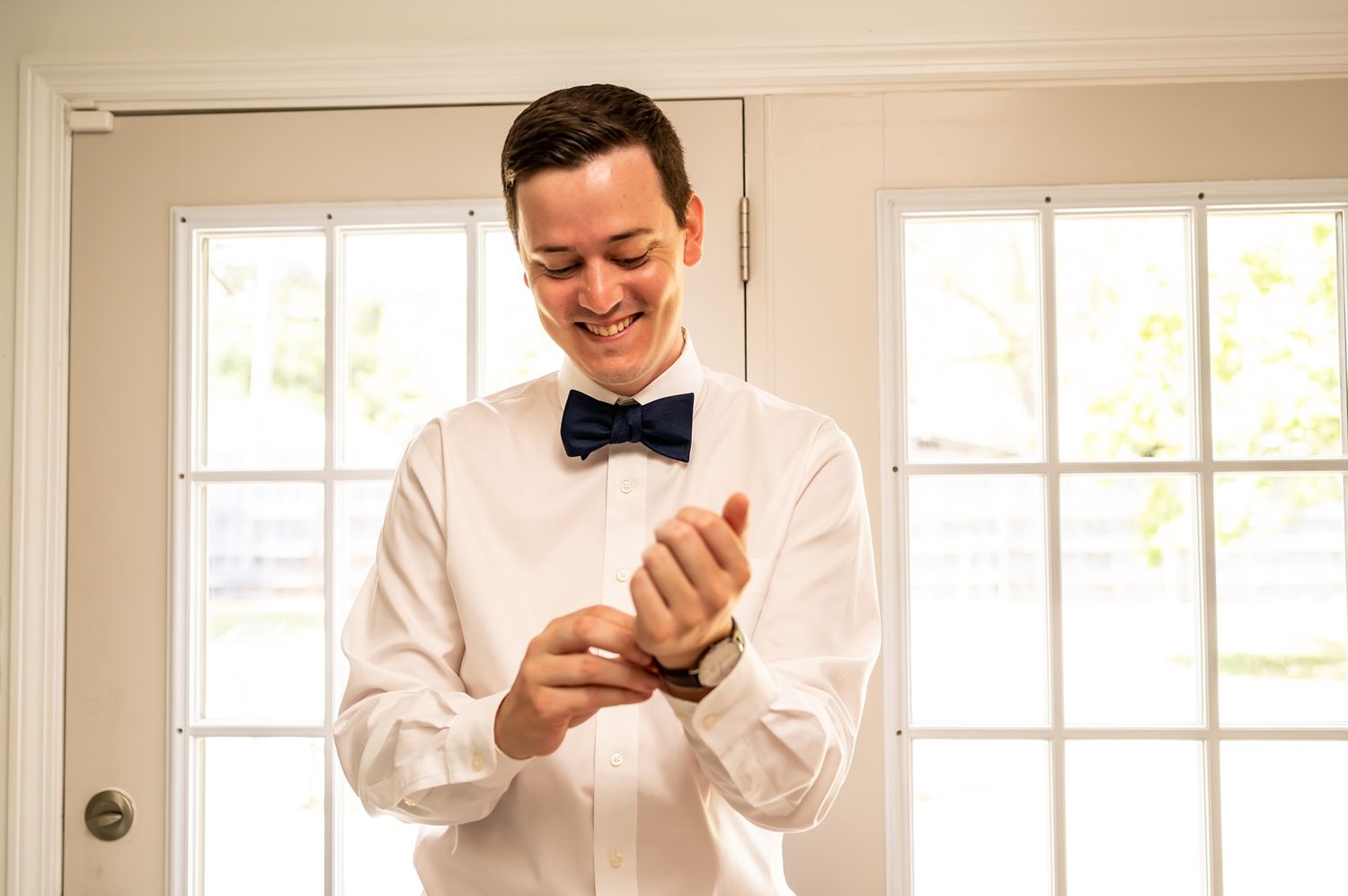 A groom wearing a white shirt and bowtie smiles while looking at his phone in a bright room with windows.
