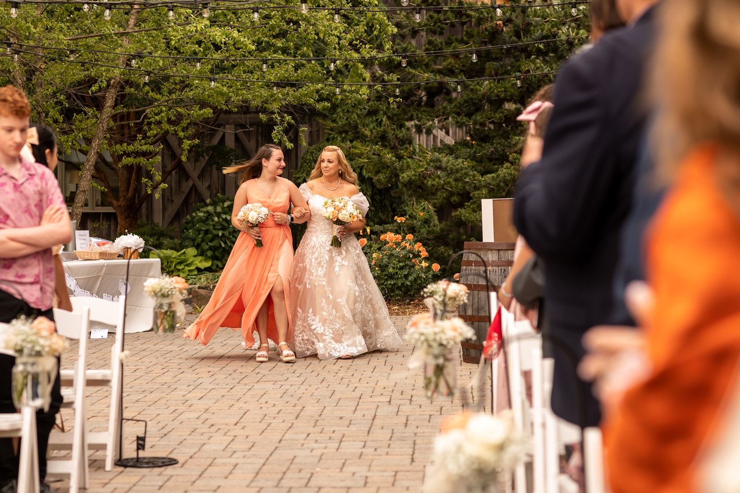 Wedding guests with peach and coral colored dresses sit in white chairs at an outdoor garden ceremony.