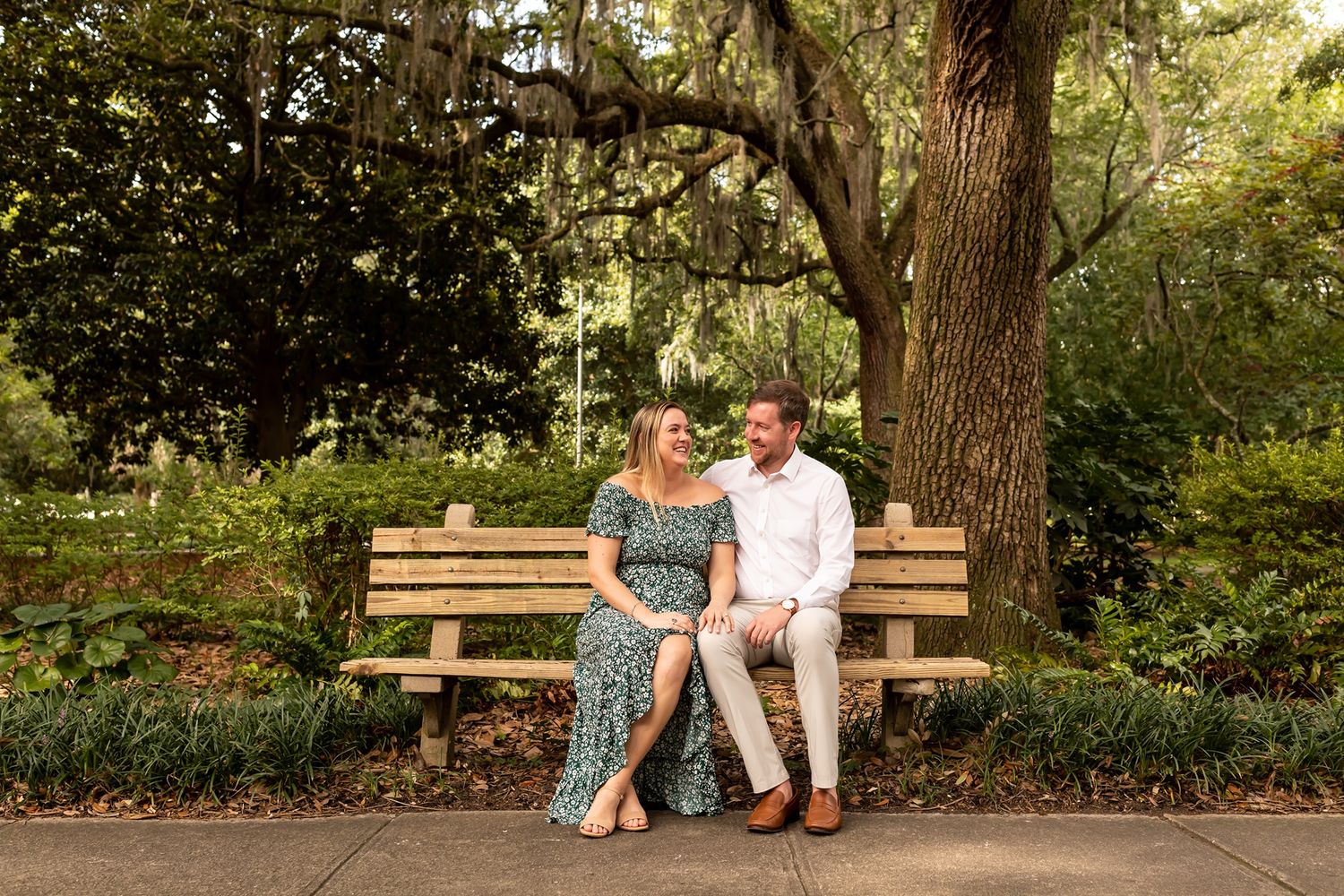 A couple sitting together on a wooden park bench surrounded by trees during a romantic outdoor photoshoot.