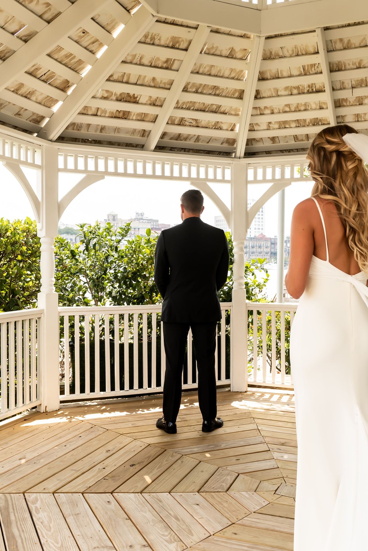 A couple stands in a white wooden gazebo overlooking a garden during what appears to be a wedding ceremony.