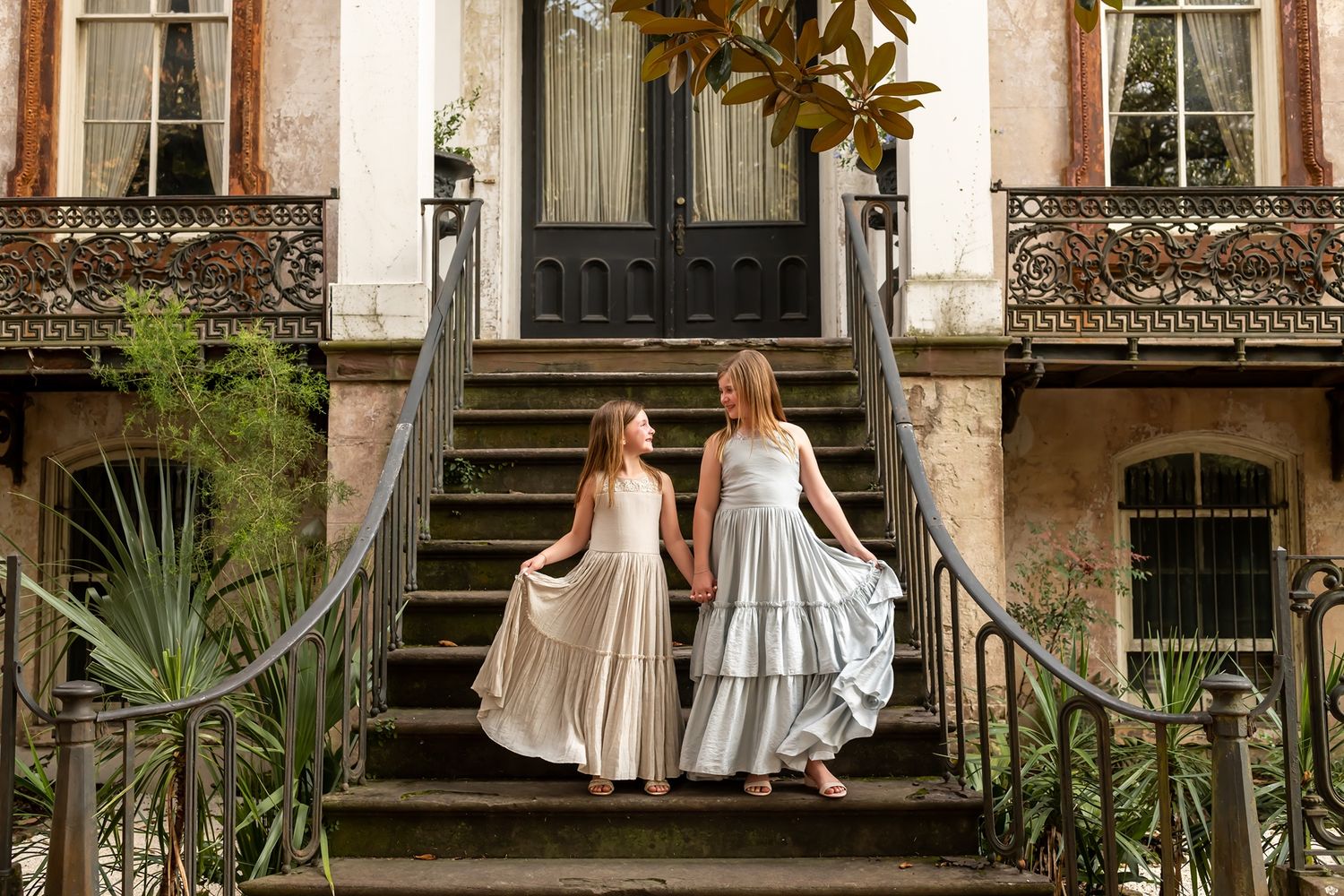 Two people in flowing dresses ascend ornate steps of a historic building with wrought iron railings and palm plants.