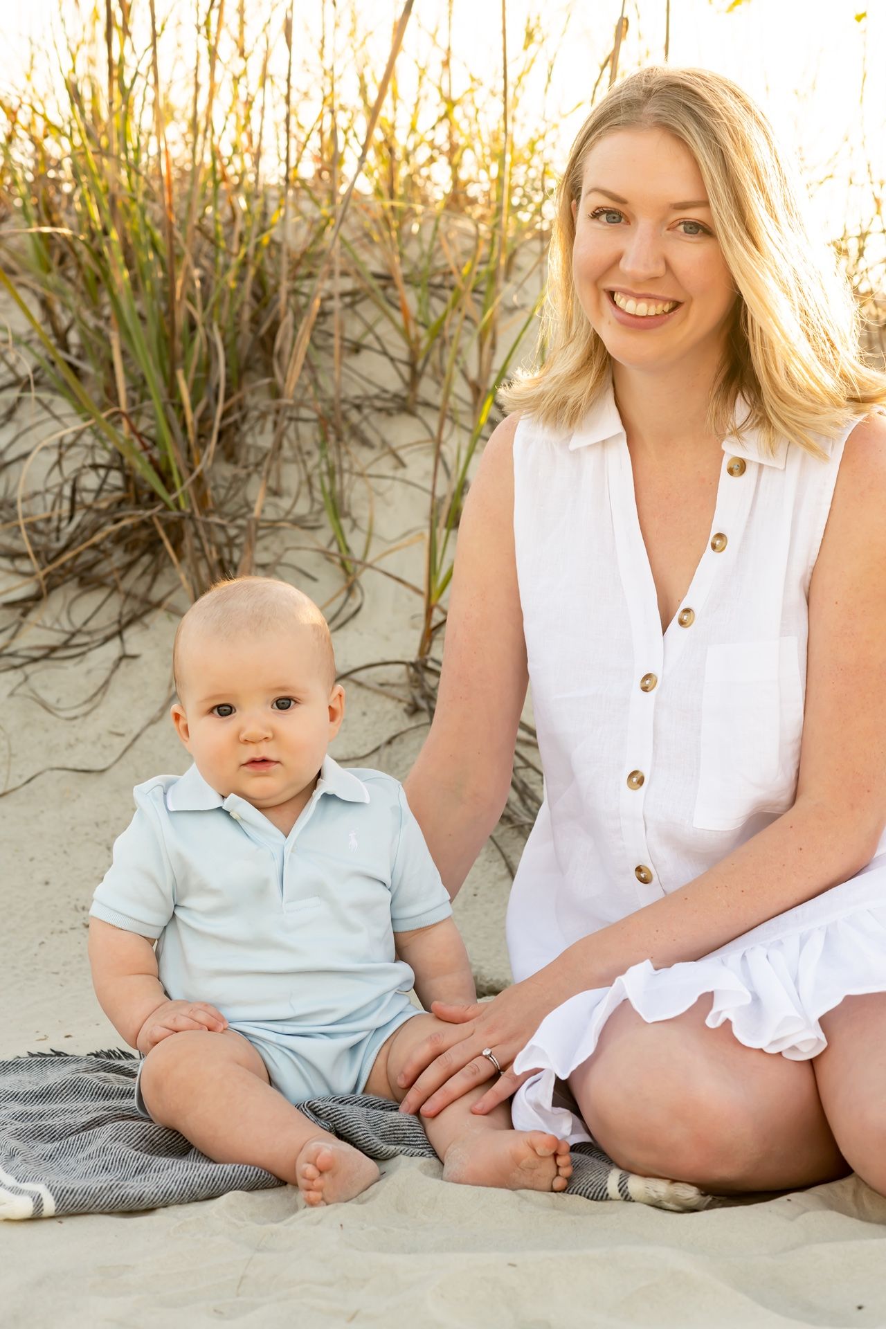 Parent and baby wearing light colored clothing sit together on blanket at beach with sand dunes in background during sunset.