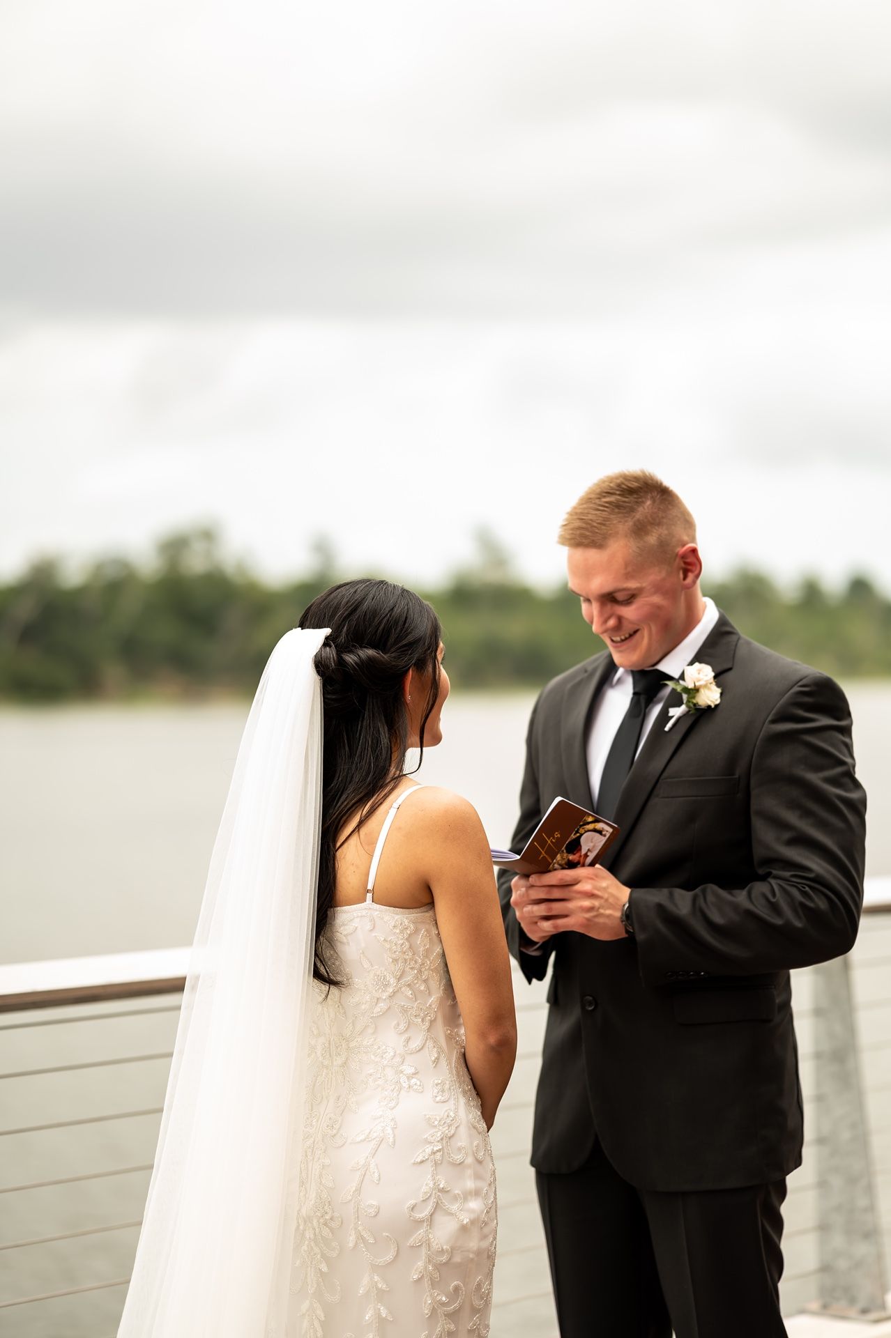 A newlywed couple stands on a waterfront deck while reading their wedding vows together.