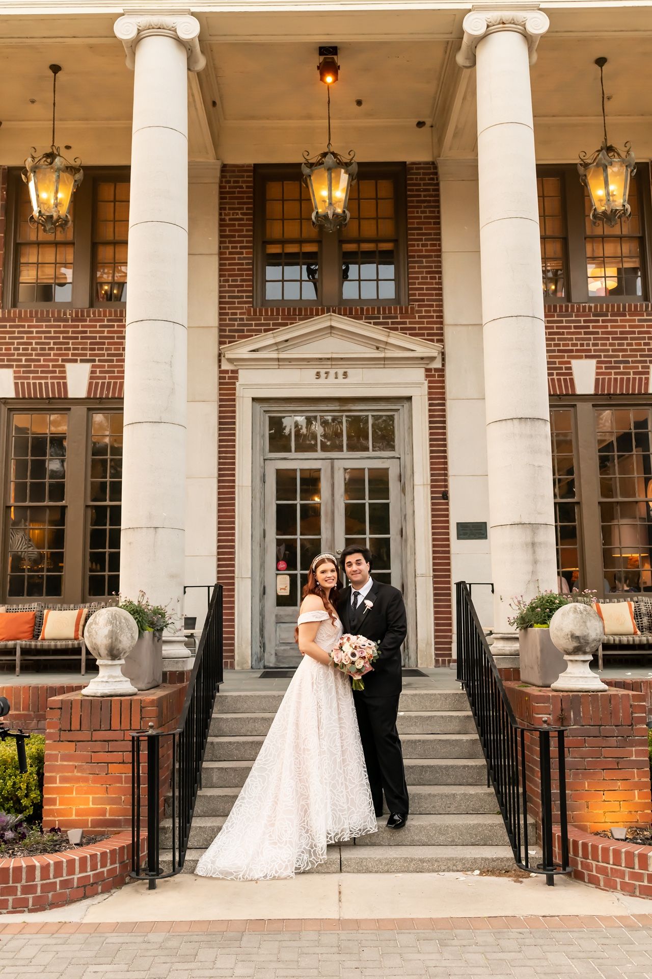 A couple poses on the steps of an elegant brick building with white columns and hanging lanterns for a wedding photo.