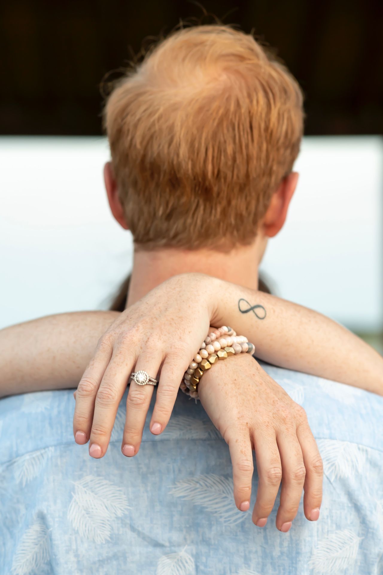 Close-up of hands wearing beaded bracelets resting on shoulders with a small infinity symbol tattoo visible.