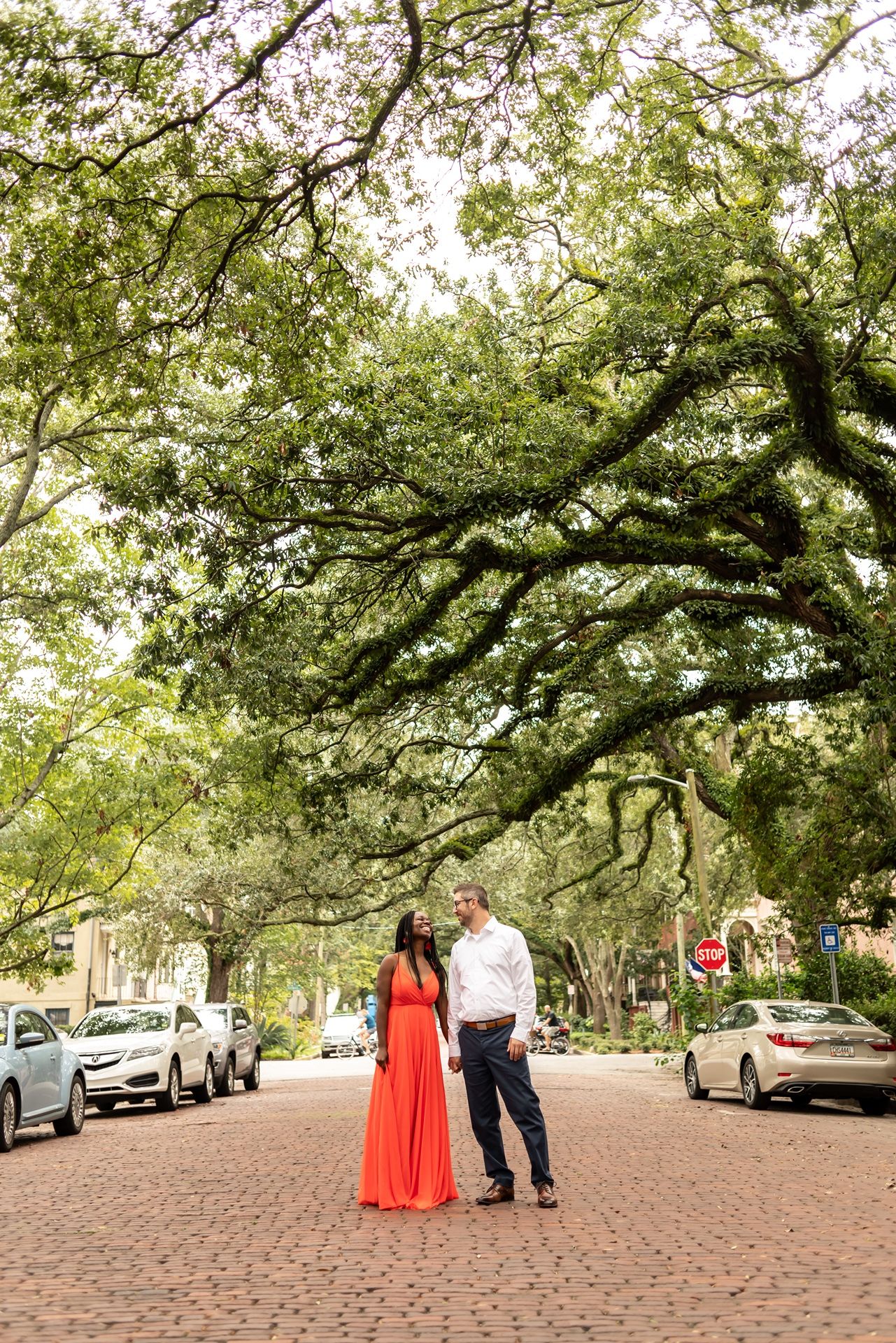 A couple in formal attire stands beneath majestic oak trees with hanging moss on a cobblestone street.