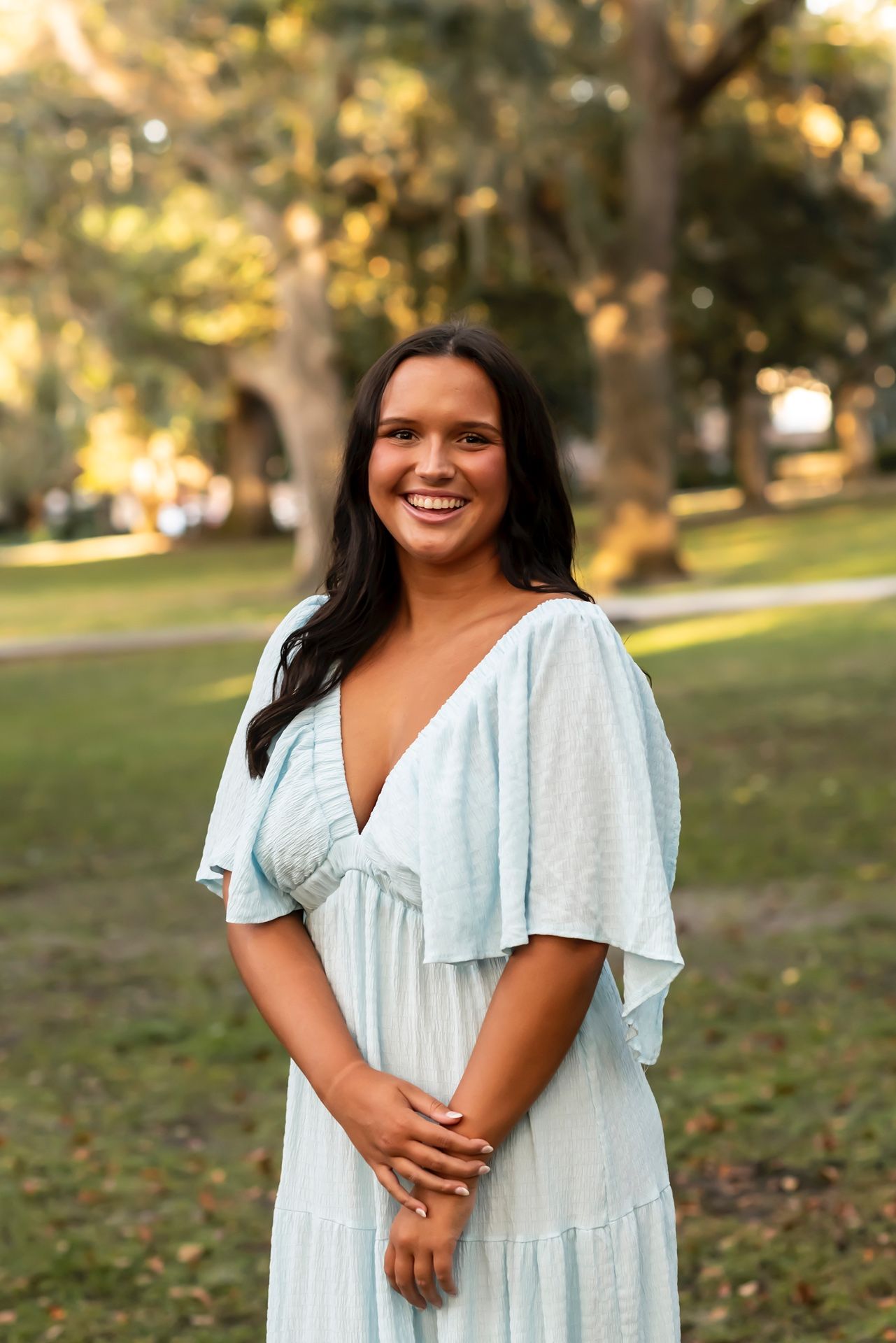 A graceful person in a light blue dress smiles warmly while standing in a sunny park setting.