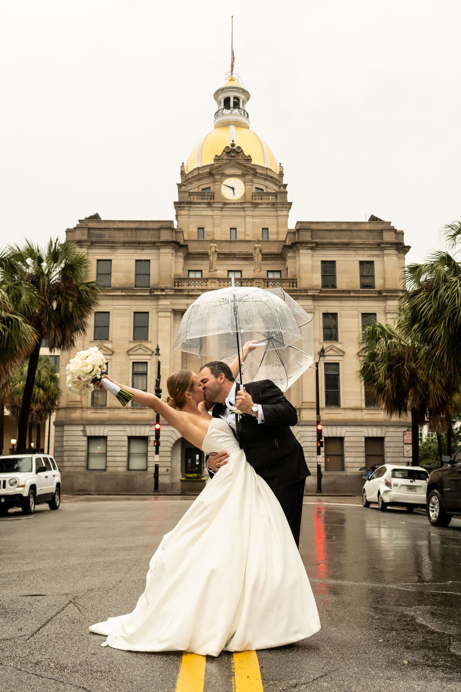 A couple with umbrella share a romantic moment outside a historic courthouse building with palm trees on a rainy day.