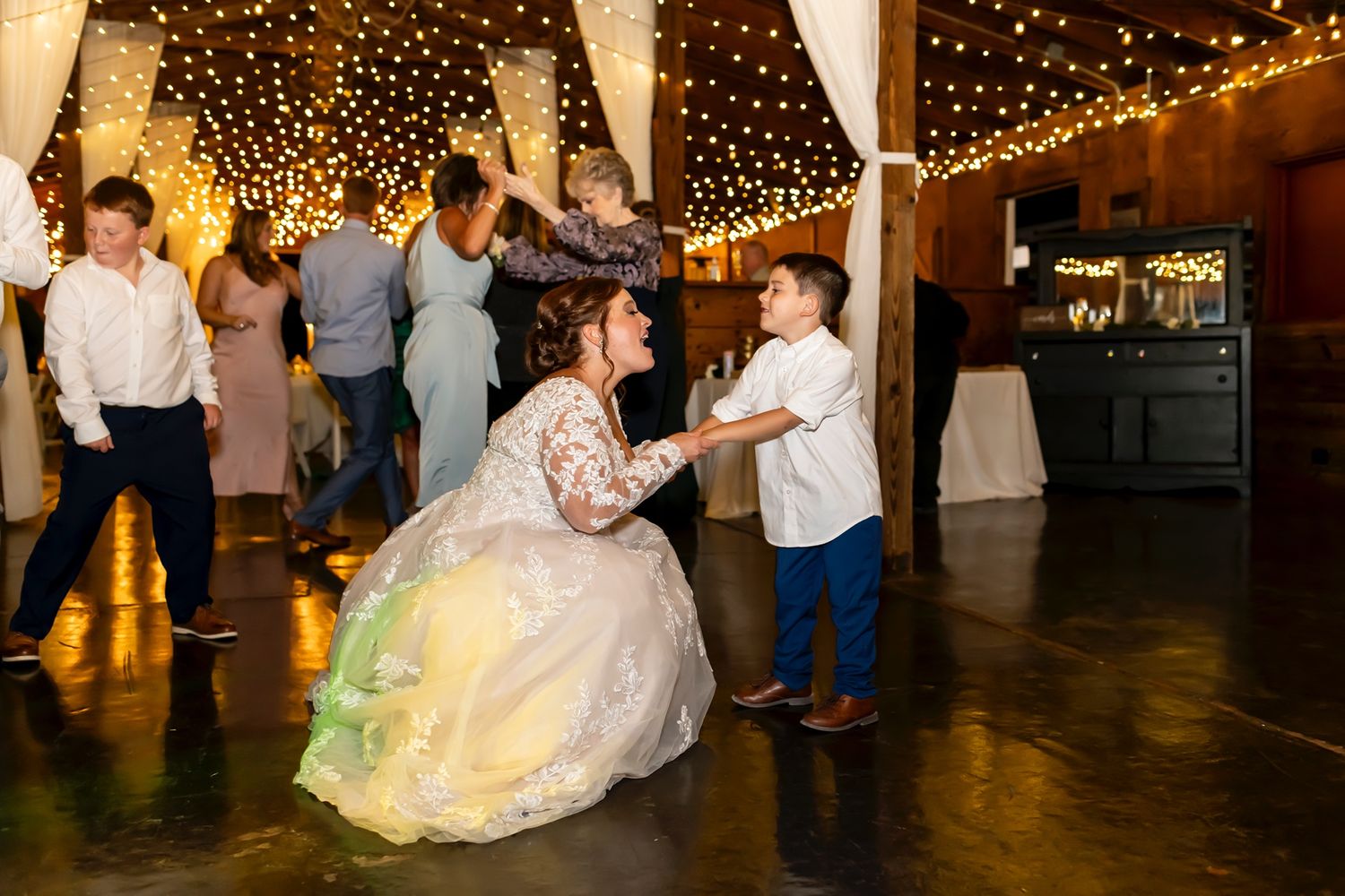A person in a white gown dances with a young child under twinkling string lights at a wedding reception.