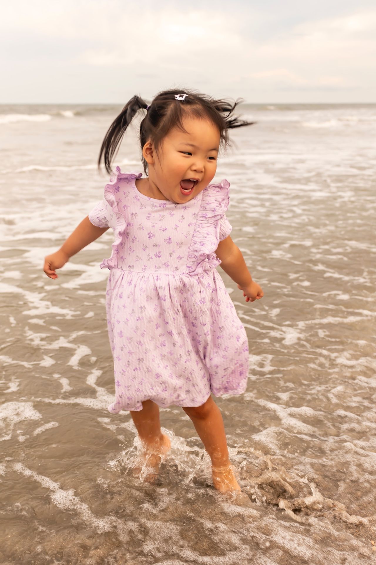 A joyful child wearing a pink polka dot dress splashes and plays in the ocean waves at the beach during a sunny day.