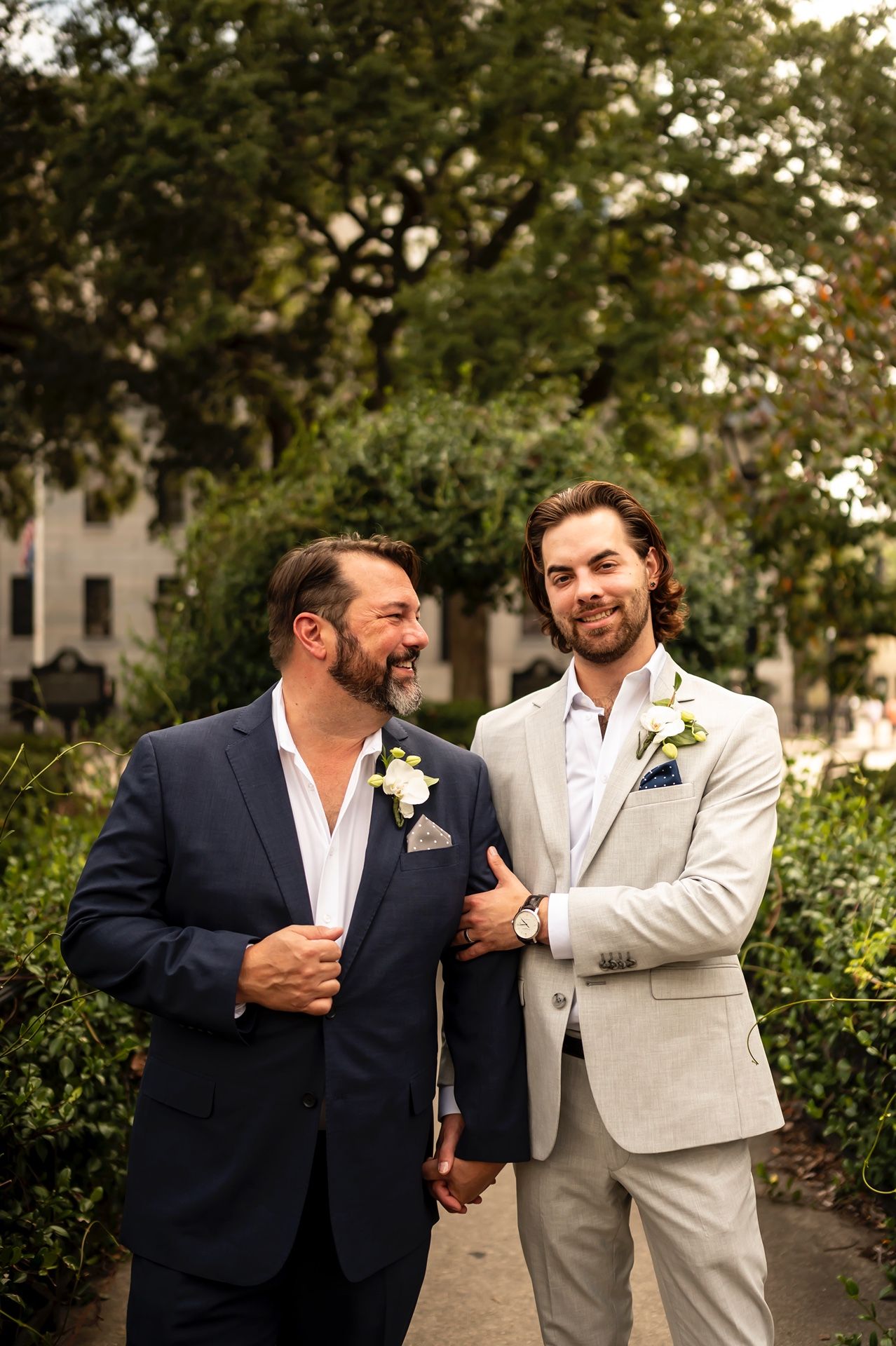 Two people in suits stand together on a garden path, wearing matching boutonnieres and sharing a joyful moment.