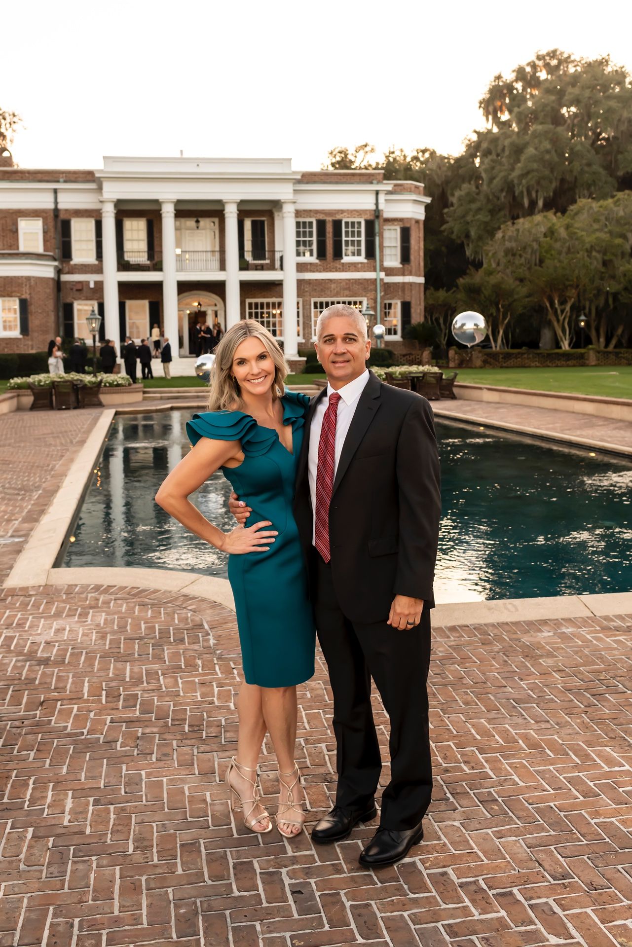 Elegant couple poses by a swimming pool in front of a grand colonial-style mansion at sunset.