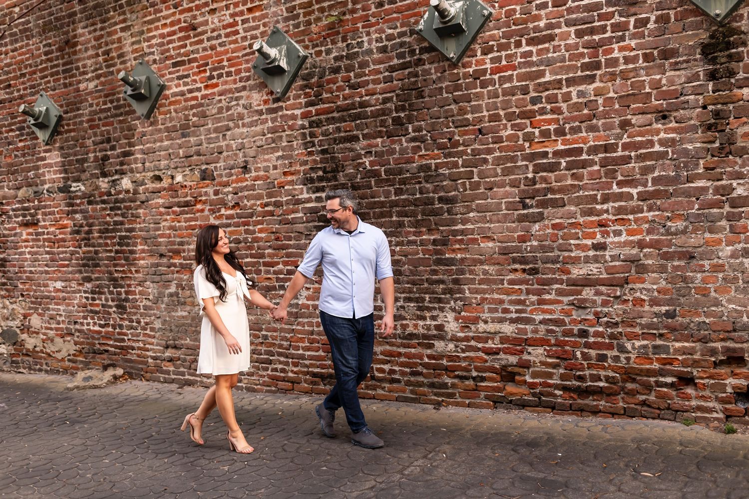 Two people holding hands while walking along an old brick wall with industrial light fixtures mounted above.